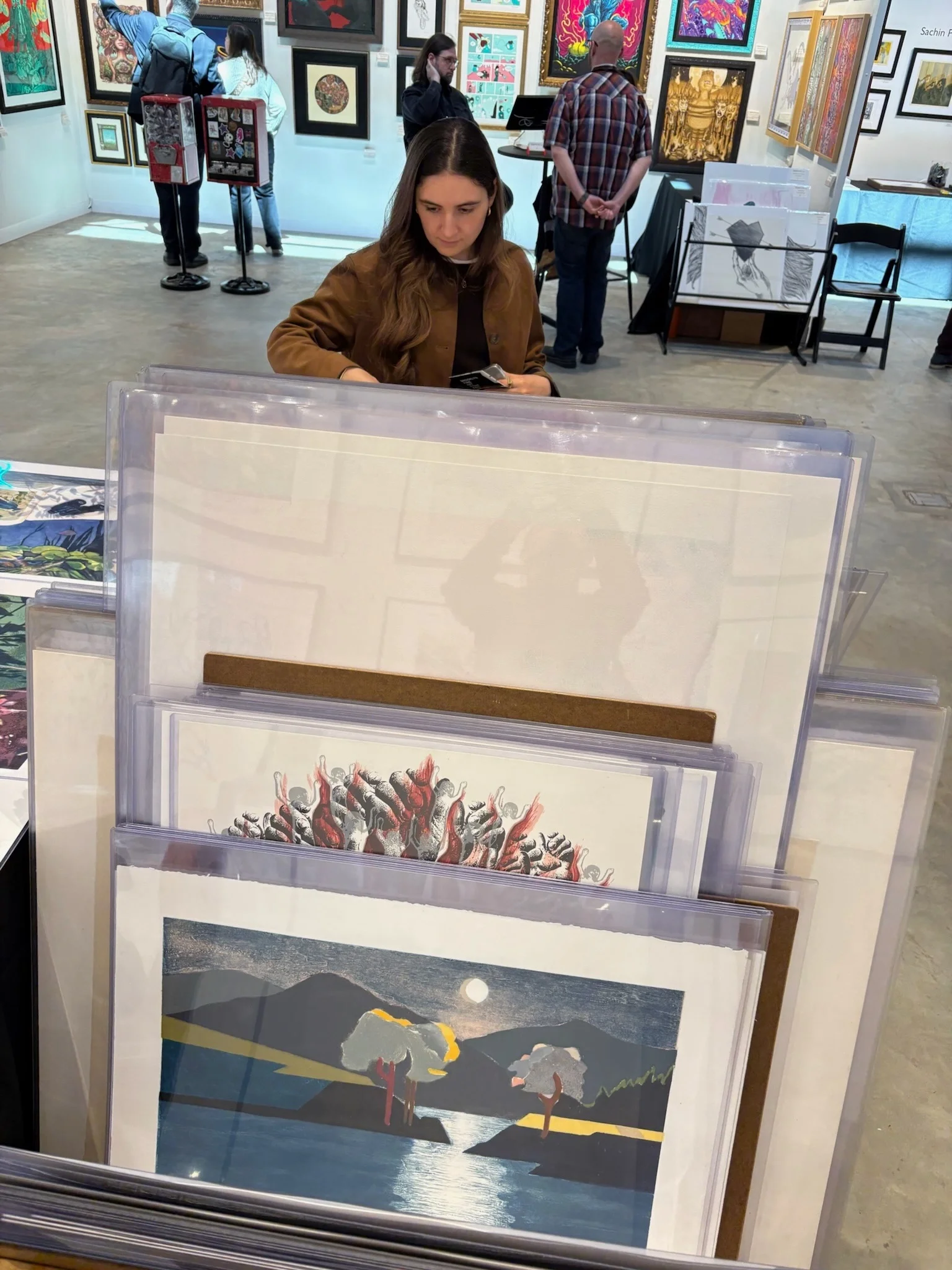 People viewing various colorful artworks in an art gallery, with one woman sitting in the foreground looking at her phone and a display of art prints in front.
