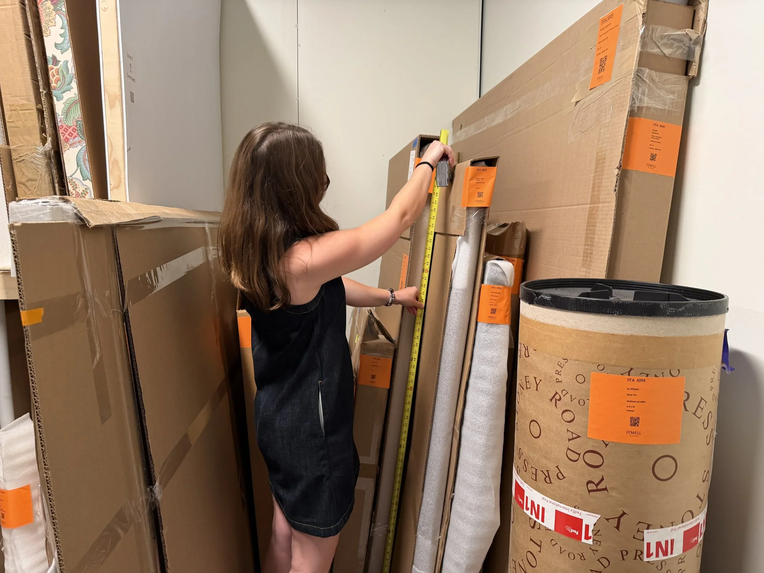 A woman with brown hair measuring a large cardboard box in a storage room or warehouse. Several other boxes are stacked around her.