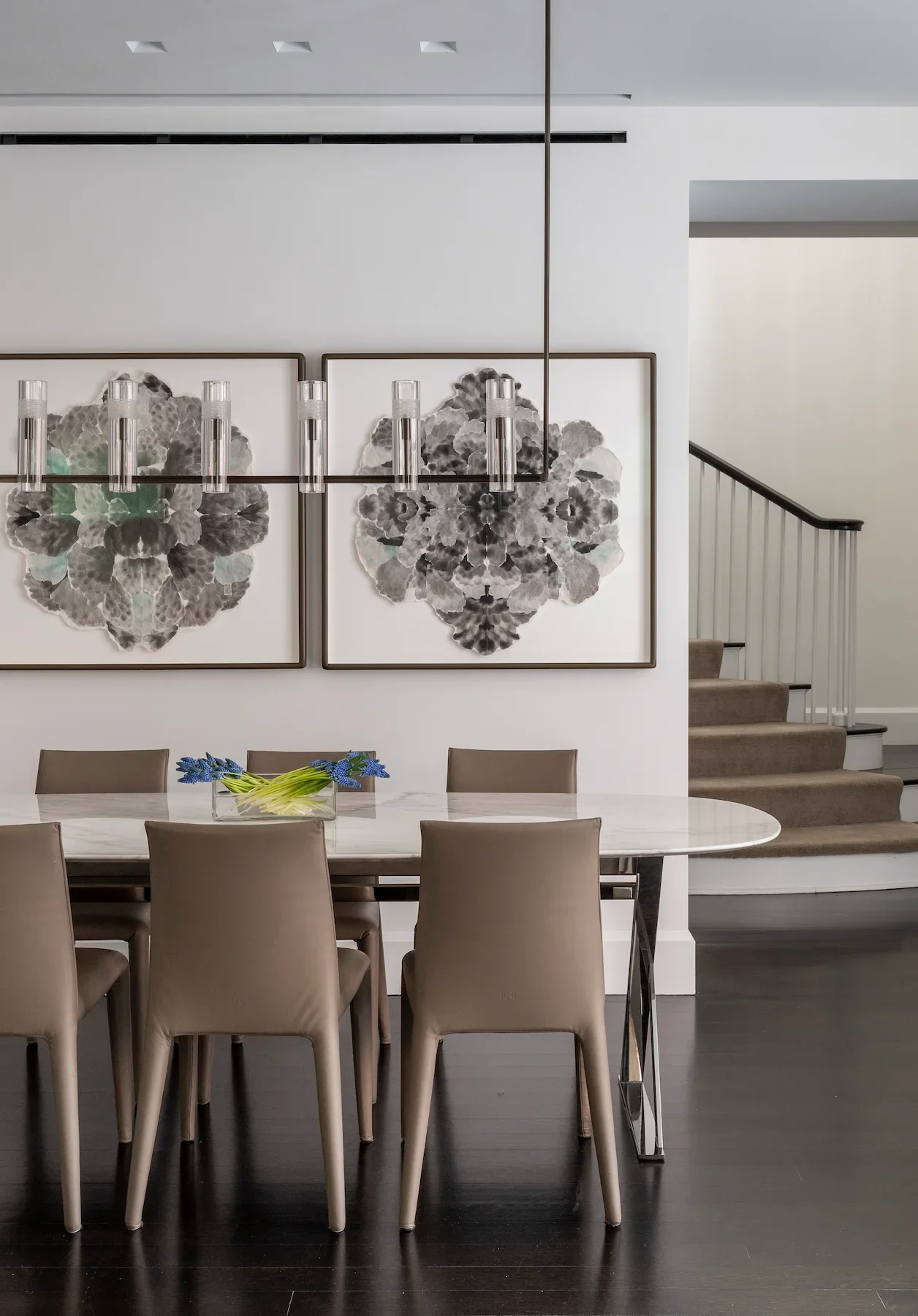 Modern dining room with a marble table, beige chairs, a floral centerpiece, and two abstract black and white art pieces on the wall. A stairway with beige carpeting and a black handrail is visible in the background.