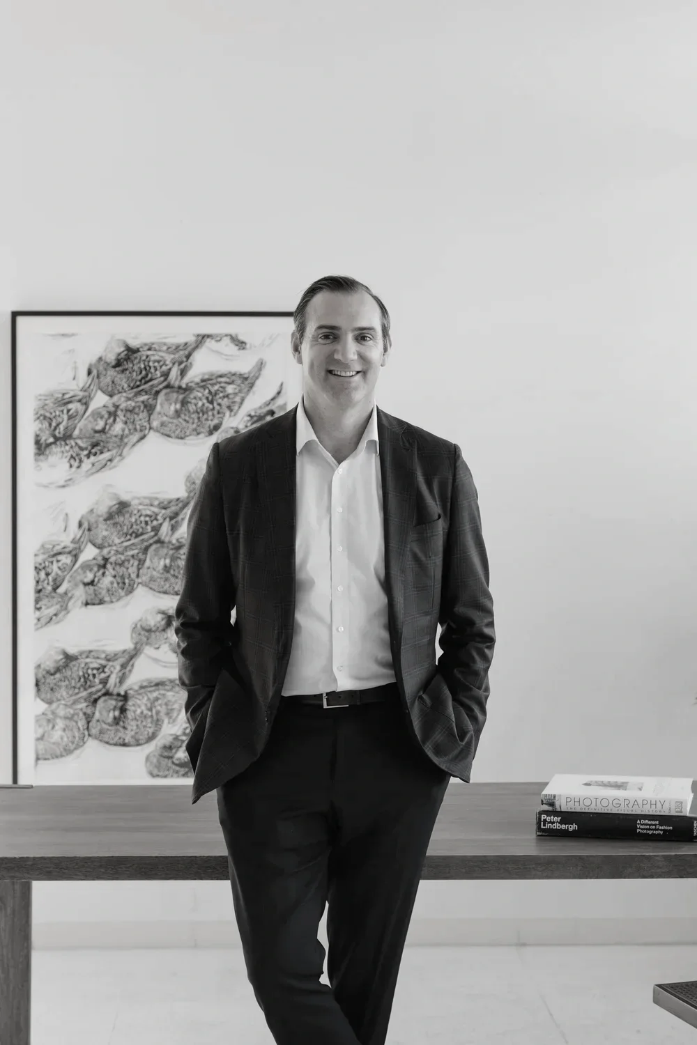 A man in formal attire standing in front of a table with books on photography, smiling at the camera. Behind him is a framed artwork of fish on the wall.