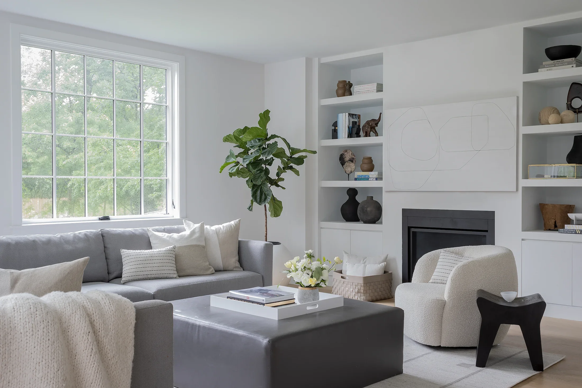 A modern living room with a large window showing green trees outside. There is a light gray sofa with beige and light-colored pillows, a white coffee table with a floral arrangement, and white built-in shelves with decorative objects and books. A green potted plant is positioned near the window, and a cream-colored armchair is in the foreground.