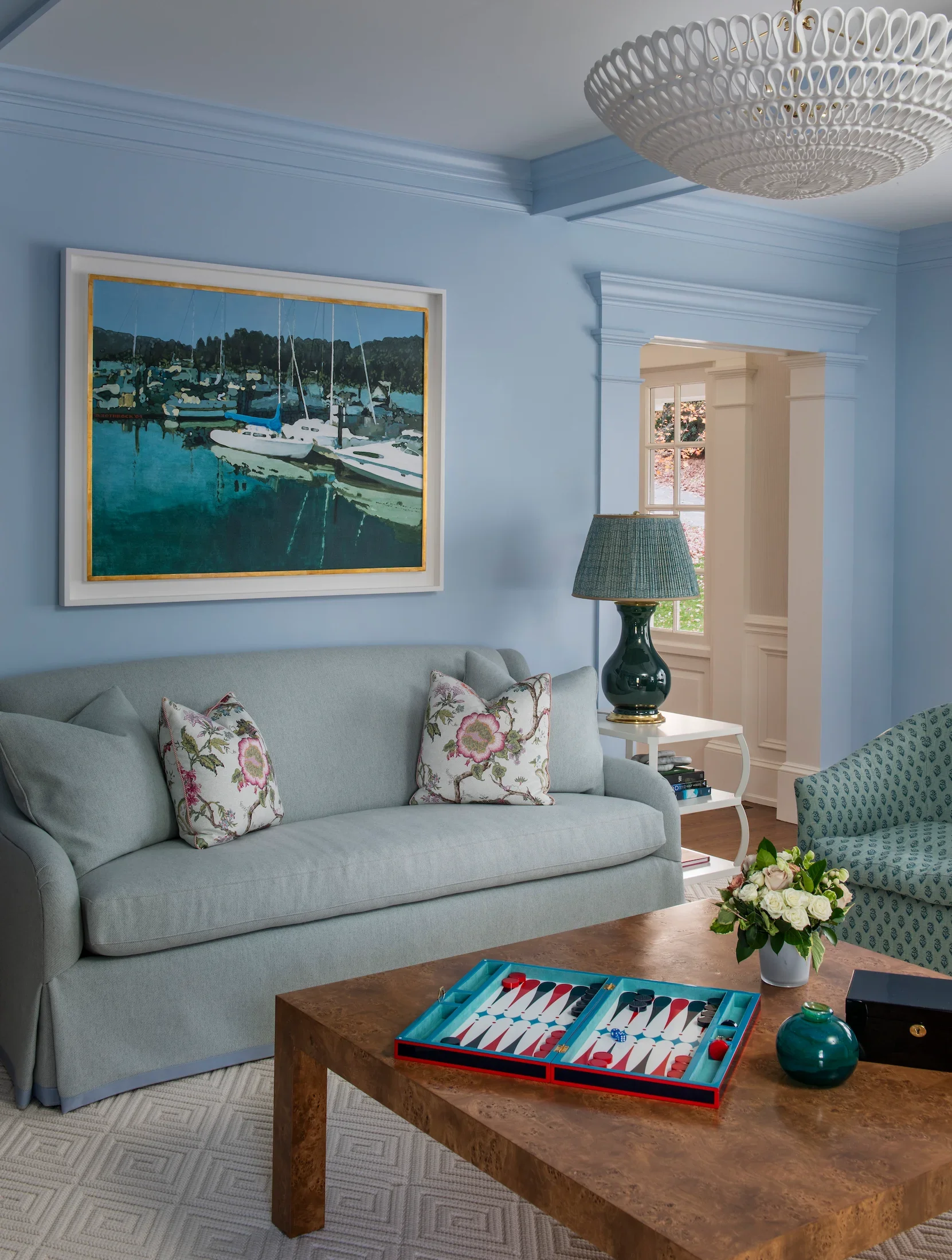 A living room with a light blue sofa with floral pillows, a wooden coffee table with a backgammon game set, a white side table with a green lamp, a framed painting of sailboats on water, a window with a view of greenery, and a ceiling light fixture.