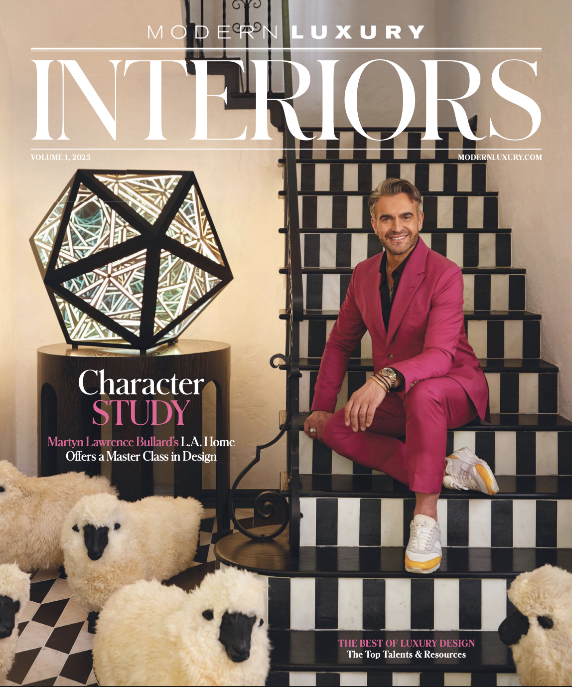 Cover of Modern Luxury Interiors magazine featuring a man sitting on black-and-white checkered stairs, wearing a bright pink suit and white sneakers, surrounded by sheep plush toys, and a geometric lamp on a side table.