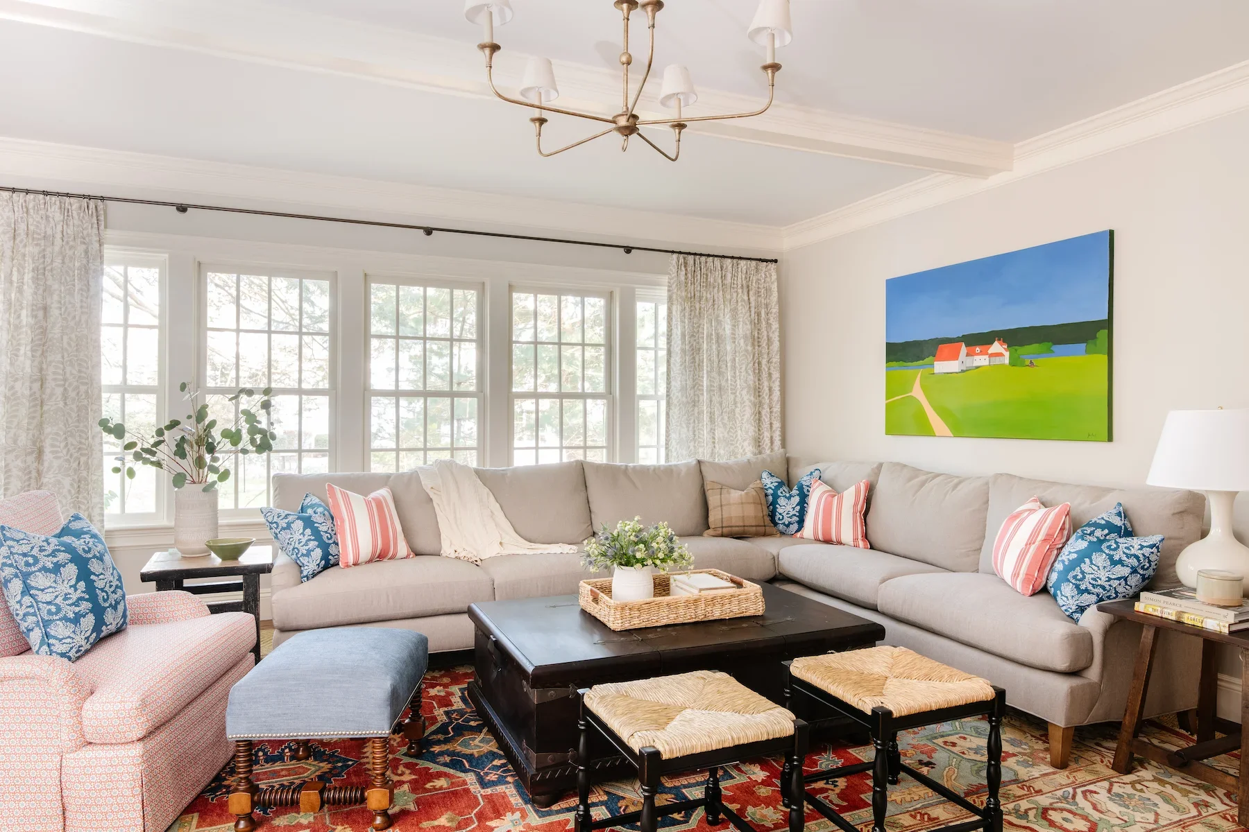 Living room with large window, beige sectional sofa with colorful pillows, a black coffee table with a wicker tray and flowers, a pink armchair, a blue ottoman, a side table with books, a white lamp, a painting of a farm scene on the wall, a chandelier, and patterned curtains.