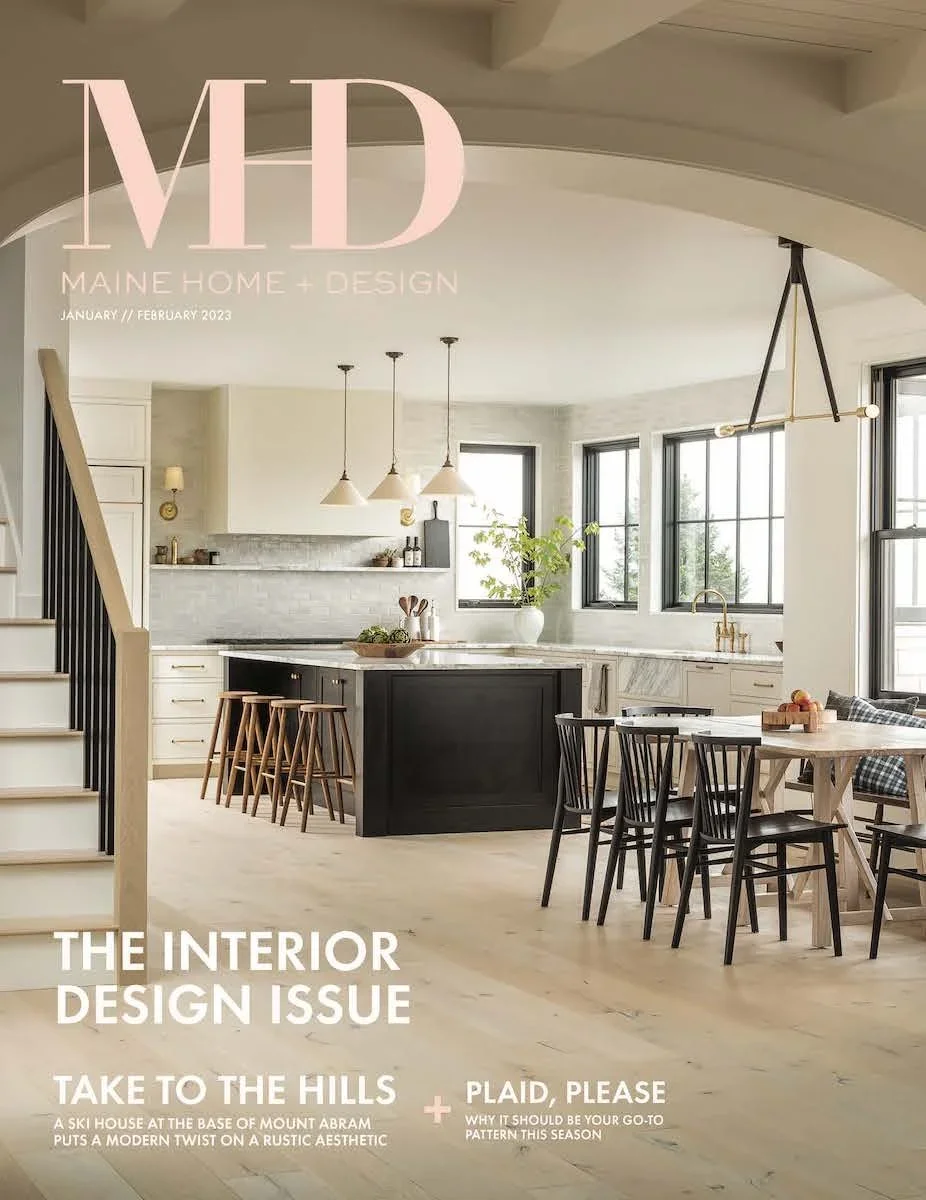 Cover of Maine Home + Design magazine featuring a modern, light-filled kitchen with white and black cabinetry, a kitchen island, a dining table with black chairs, large windows, and minimalist decor.