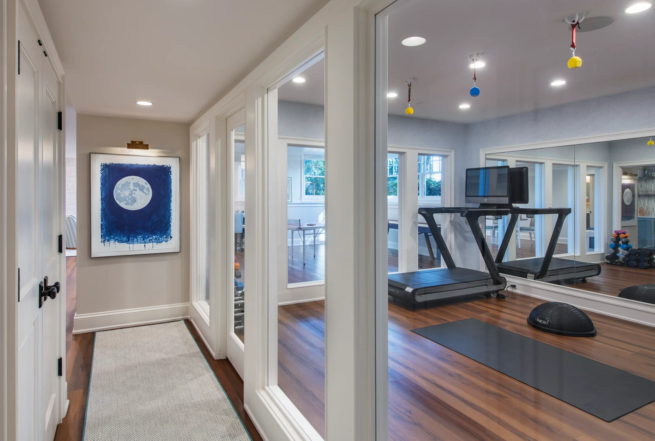 A home gym enclosed with glass walls, featuring a treadmill, a set of dumbbells, a black exercise mat, and colorful hanging decorations on the ceiling. There is a hallway with a framed art piece of a full moon on the wall.