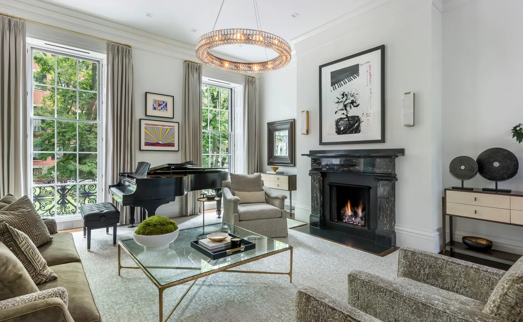 A bright living room with large windows and a view of green trees outside. The room features a black grand piano, a modern white side table, a cozy armchair with a pillow, a fireplace with a fire burning, various art pieces on the walls, and elegant decor including a chandelier and sculptures on a console table.