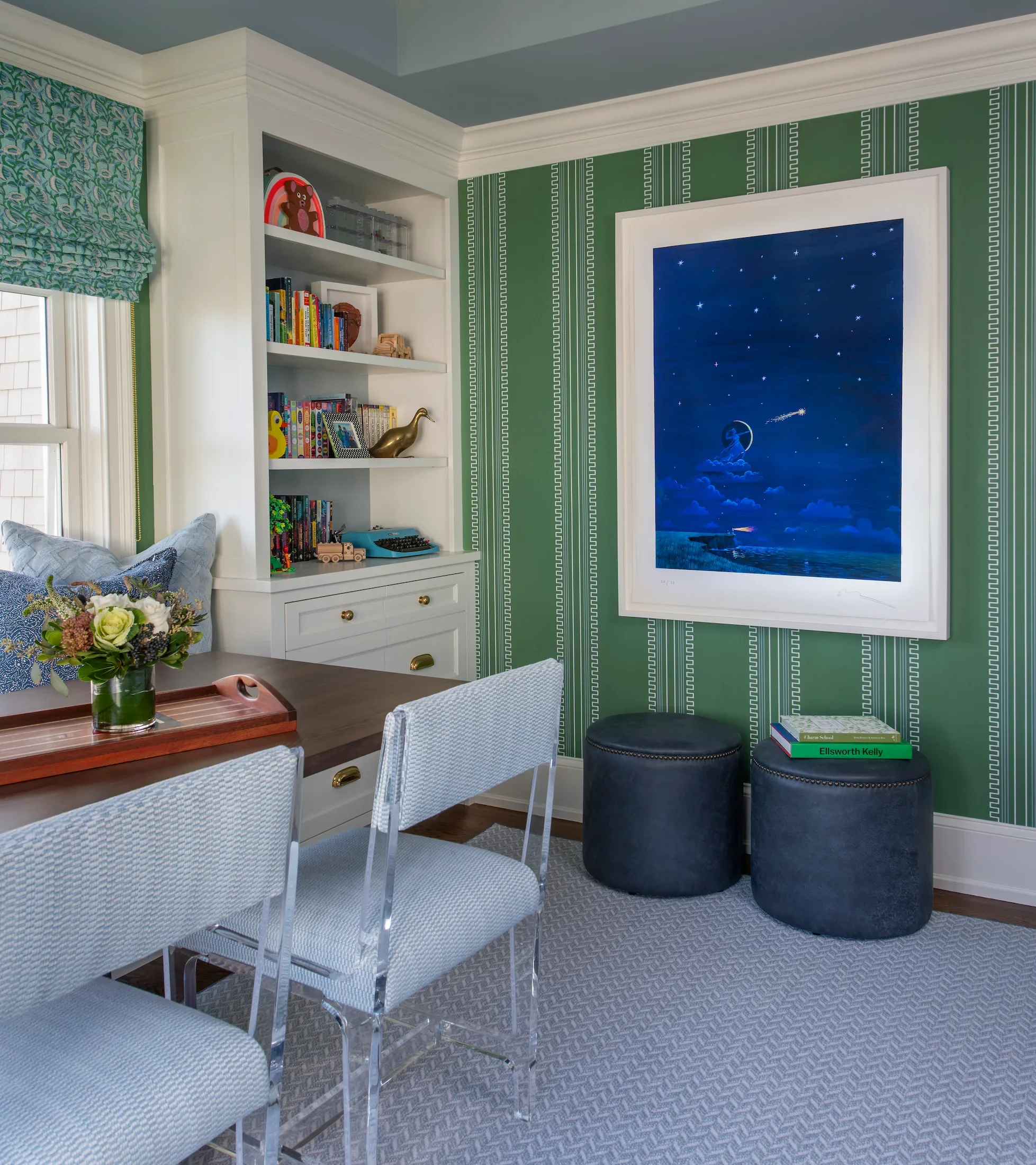 A family room with green decorative walls and a large framed painting of a night sky with stars and a moon. There is a white built-in bookshelf filled with books, small decorative items, and a picture frame. A window with a patterned green valance letting in natural light is visible on the left. A table with a flower arrangement and a tray is in the foreground, with transparent chairs around it. Two dark round ottomans are next to the wall, with a couple of books on top.