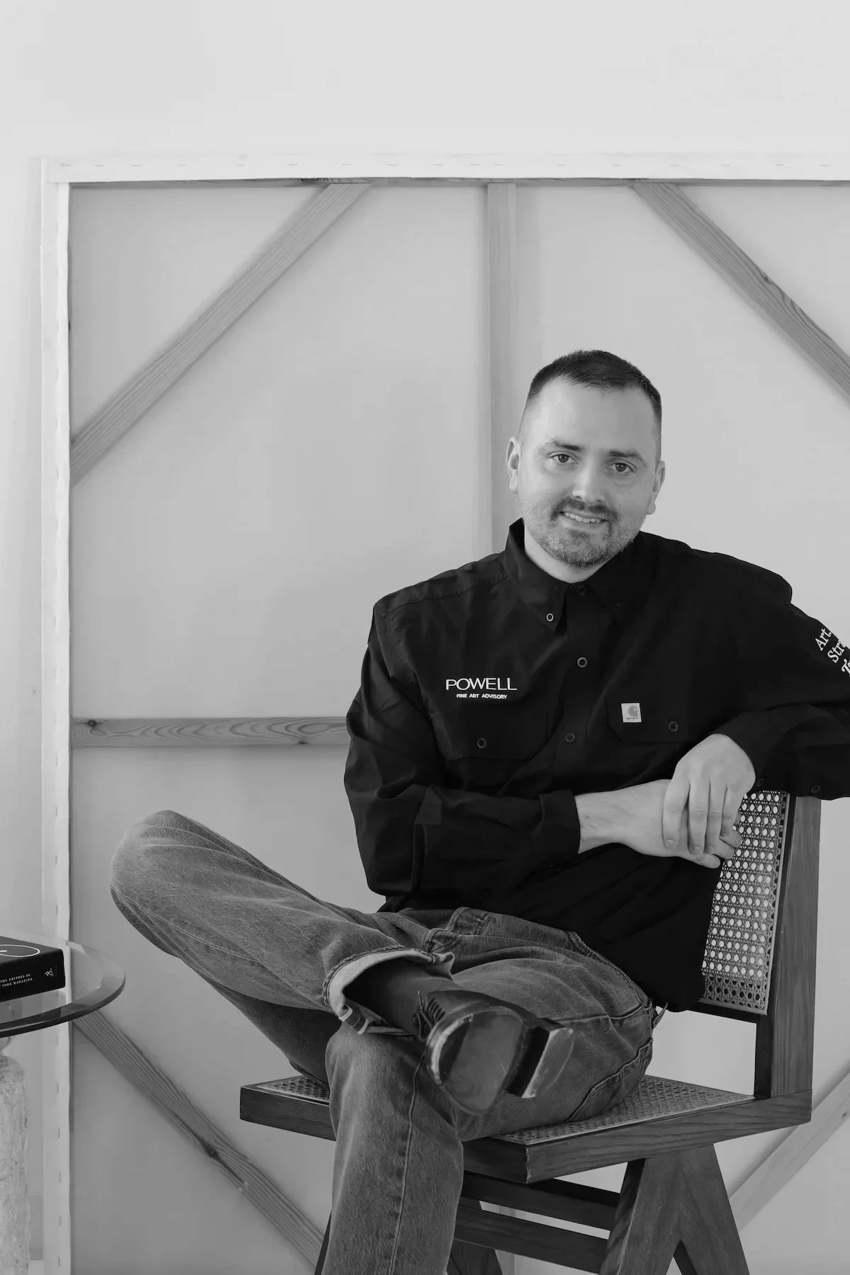 A man with short hair and a beard wearing a black work shirt with the name Powell and a patch on the sleeve, sitting on a chair with one leg crossed over the other. He is smiling at the camera, with a background of wooden framing and drywall.