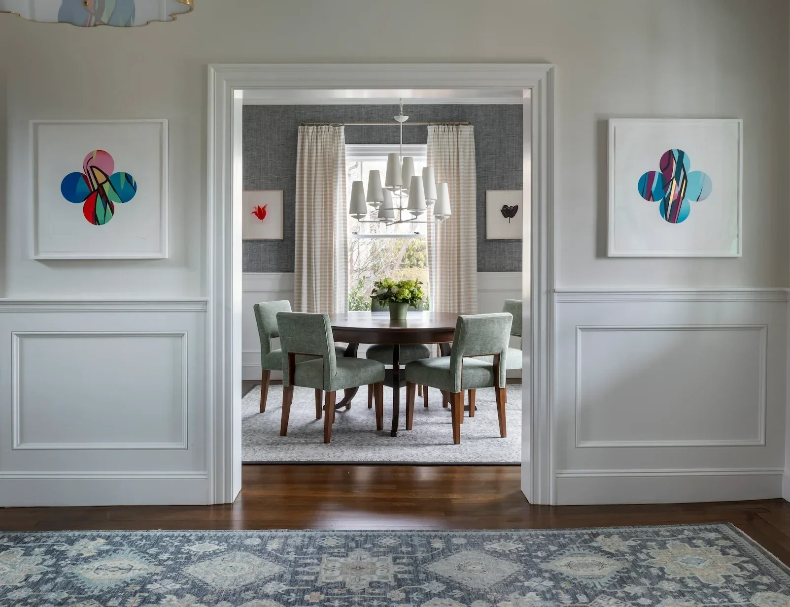 A dining room seen through an open doorway, featuring a round wooden table with a vase of flowers, surrounded by four green upholstered chairs. The back wall has a window with checkered curtains, and a modern chandelier hangs above the table. There are framed artworks on the walls.