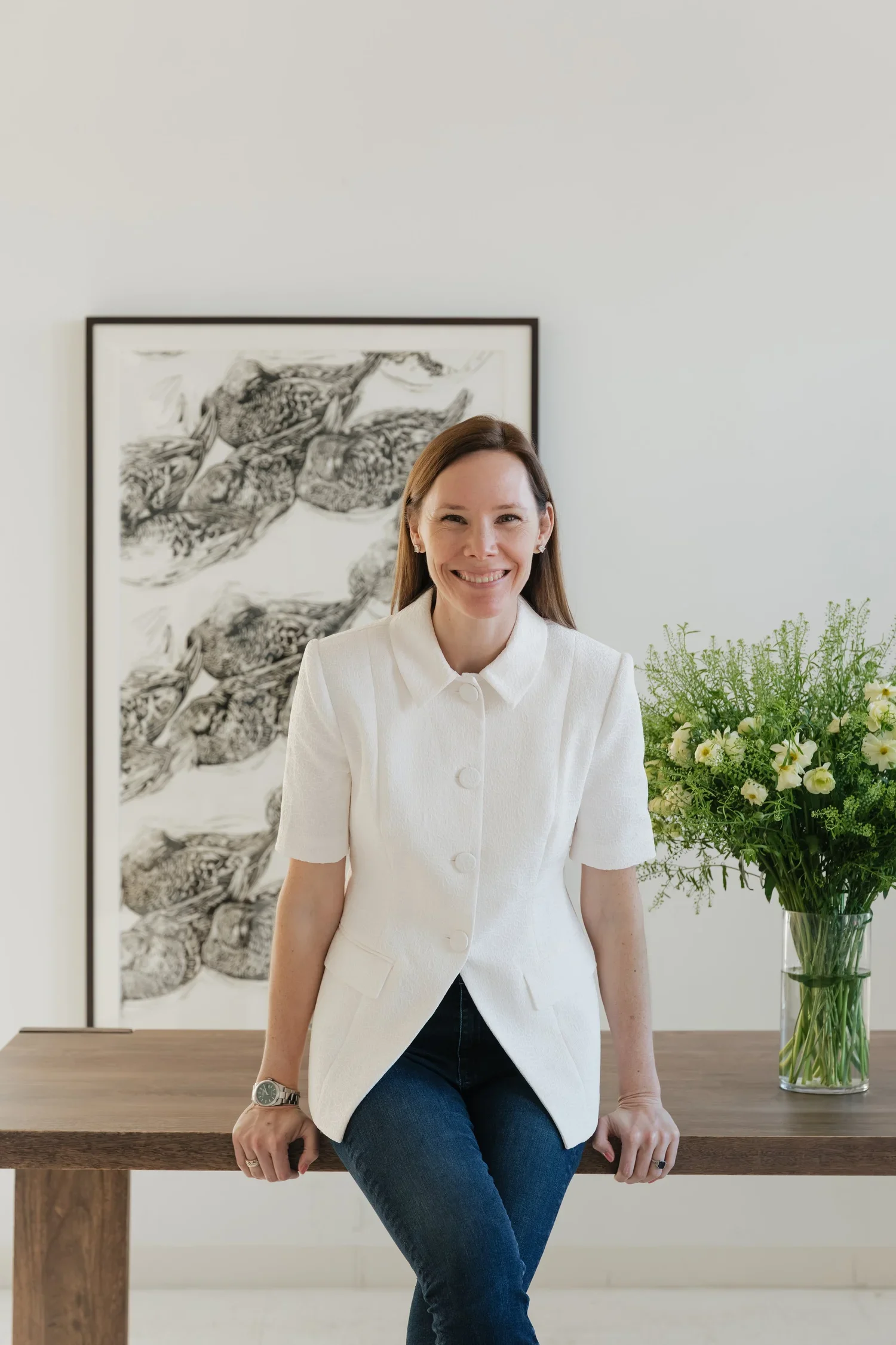 Woman sitting on a wooden table in front of a white wall with black and white artwork and a green floral arrangement nearby, wearing a white blazer and blue jeans, smiling.