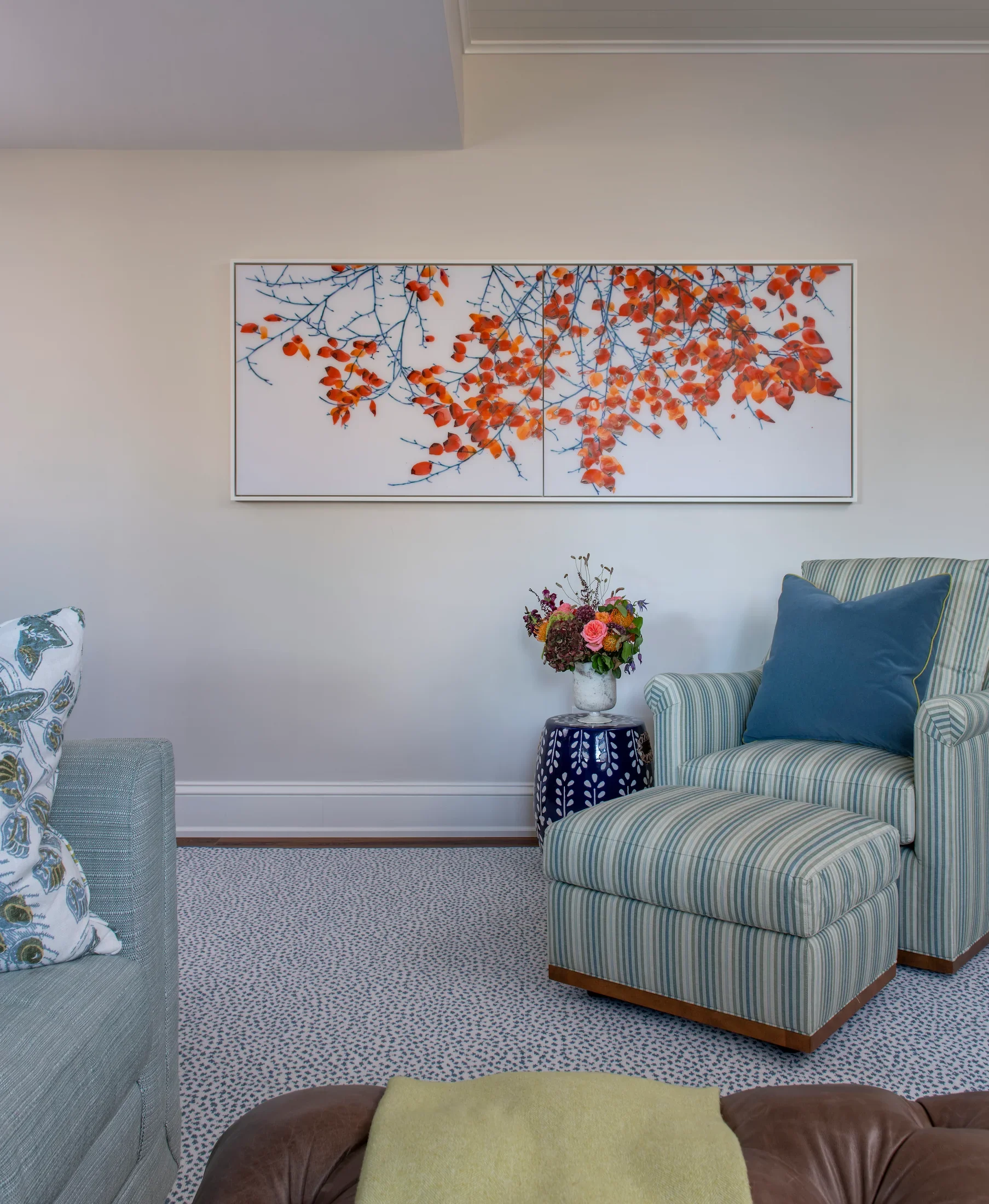 Living room with gray armchair on left, striped armchair and ottoman on right, colorful flower arrangement on small black table, large wall art of orange and blue branches, carpeted floor.