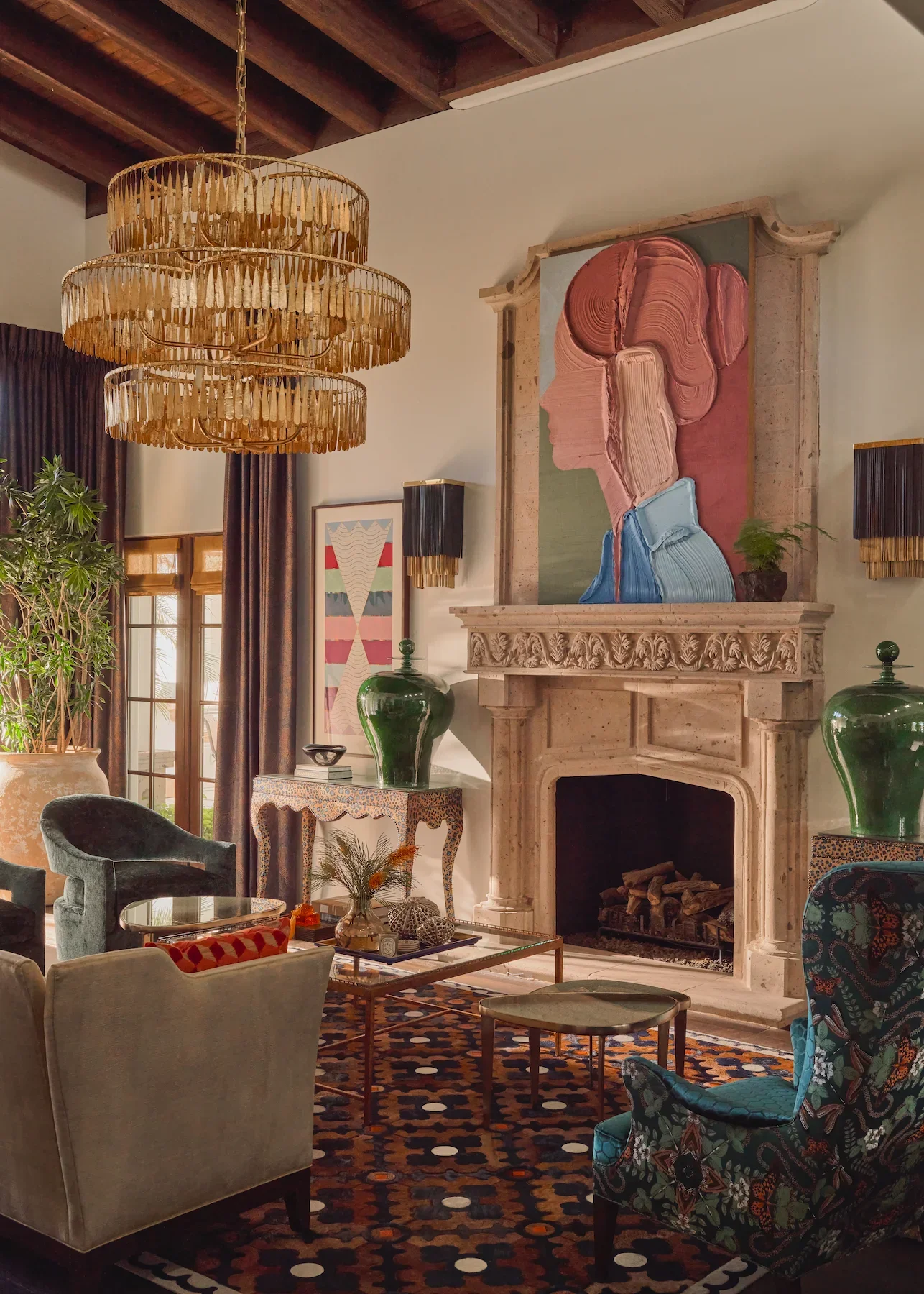 Living room with a fireplace, a large abstract portrait painting above it, velvet and patterned chairs, a glass coffee table, large green vases, a decorative rug, plants, and a chandelier.