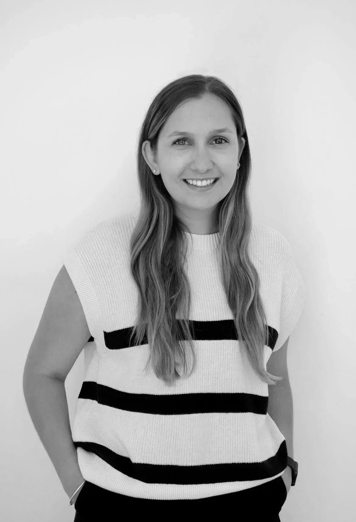Black-and-white photo of a smiling woman with long, wavy hair, wearing a striped sleeveless top, standing against a plain white background.