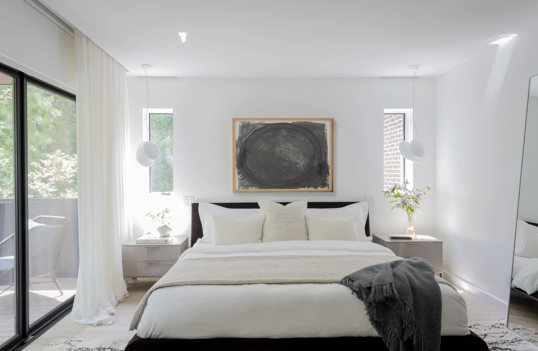 A modern bedroom with white walls, a bed with white bedding, a black headboard, and decorative pillows. Two white nightstands with books and a lamp are on either side. Above the bed is a framed abstract black and white artwork. Large windows with white curtains let in natural light, and a plant with purple flowers is next to the right window.