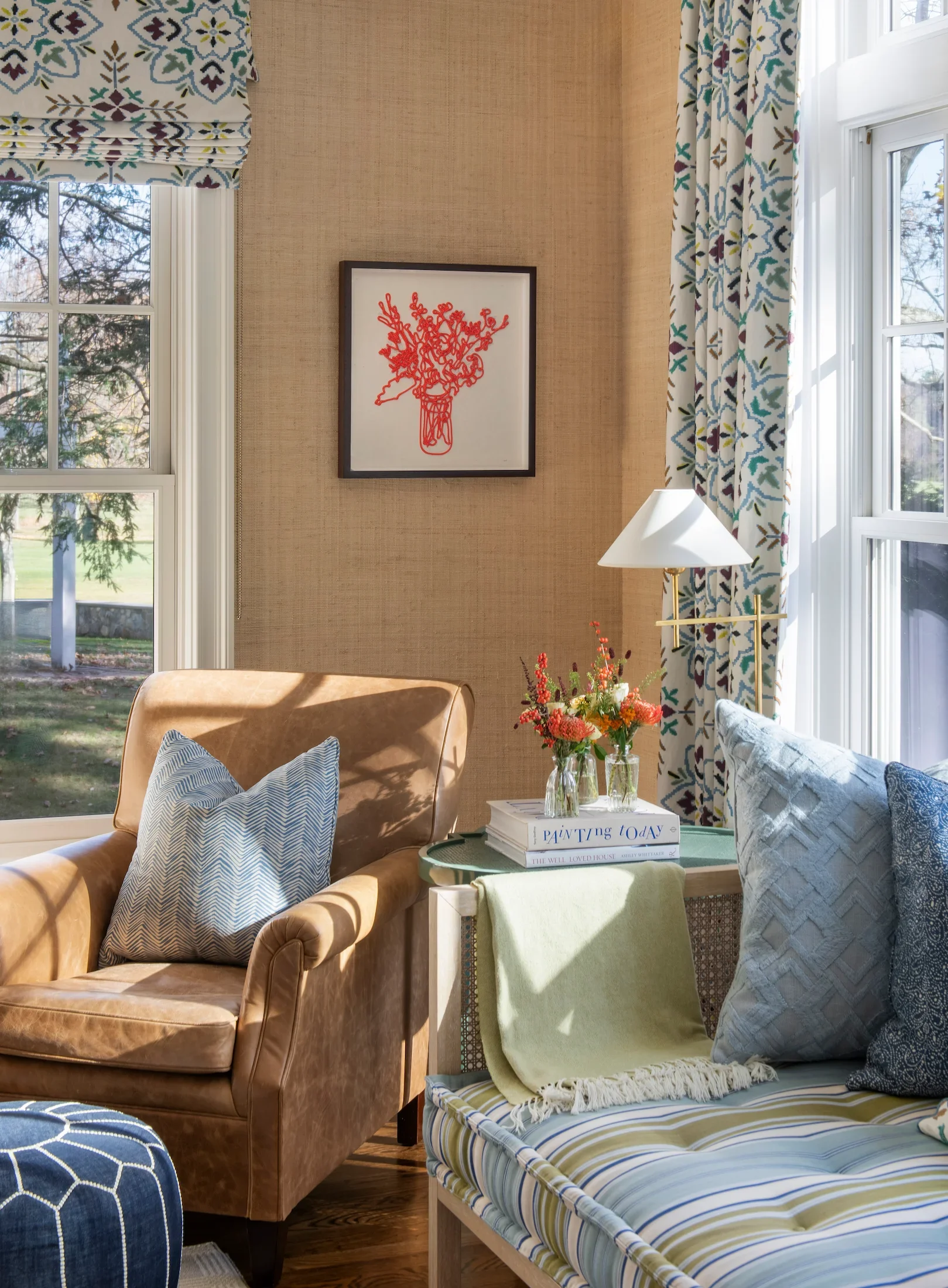 A cozy living room with beige walls, large windows with patterned curtains, a brown leather armchair with striped pillows, a side table with colorful flowers and books, and a framed red line art of a vase with flowers hanging on the wall.