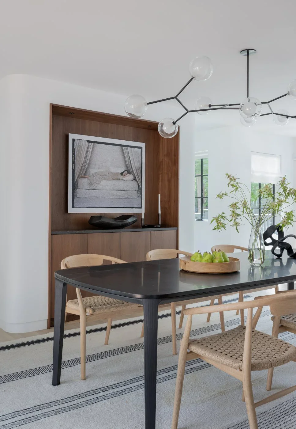 Modern dining room with black table, light wood chairs, a vase with green branches, a tray of green apples, a black sculpture, a wooden cabinet with artwork, and a contemporary chandelier.