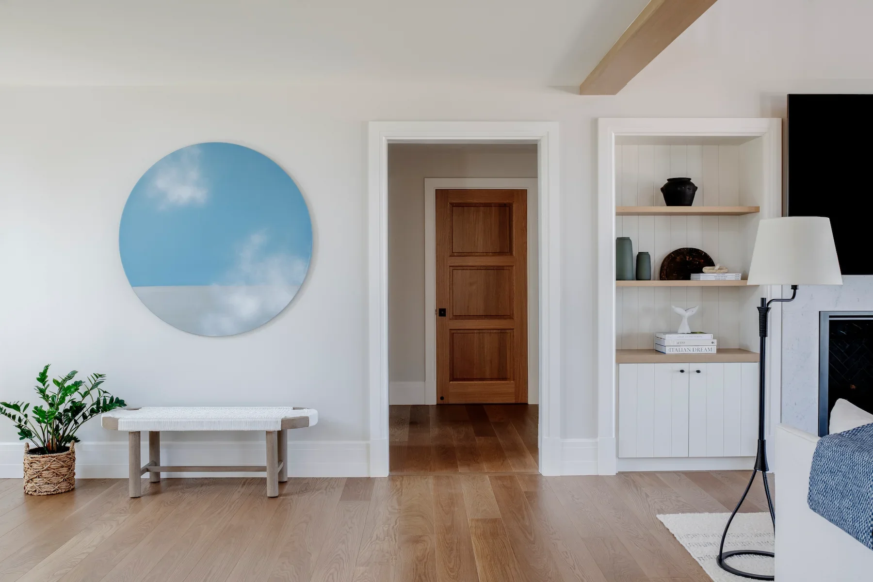 Living room with white walls, wooden floor, and minimal decor. A round mirror reflects the sky, a small bench with a plant in a woven basket, built-in shelves with vases and books, a white lamp, and partially visible sofa and fireplace.