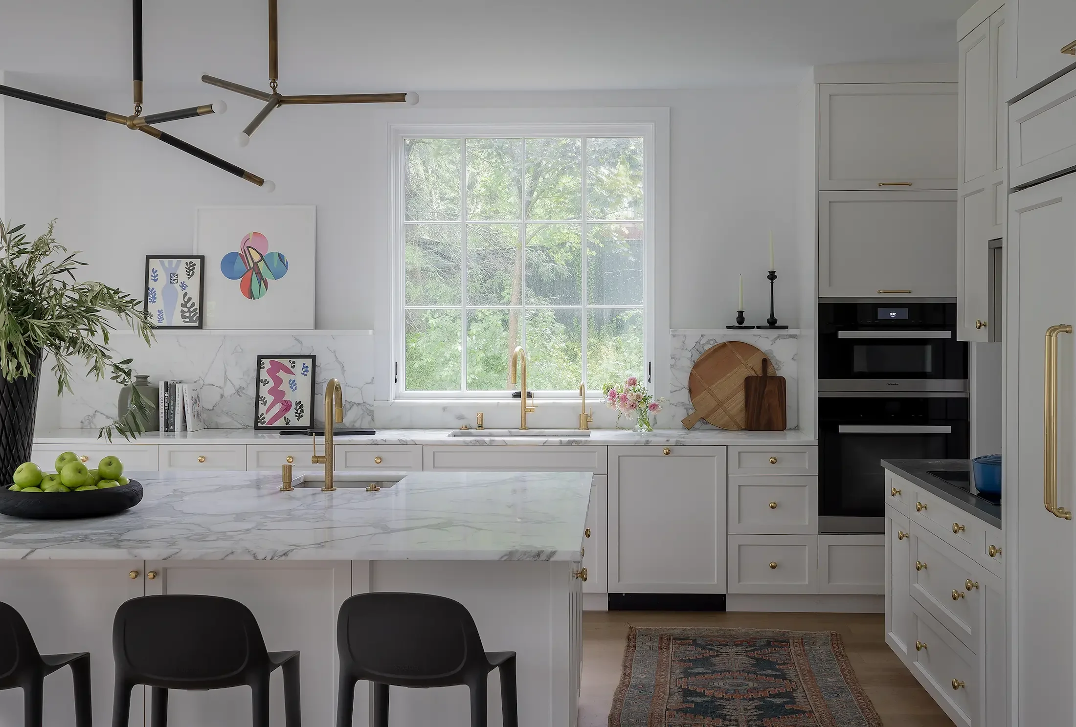 A modern white kitchen with large window, marble countertops, black chairs, and decorative art on the wall.