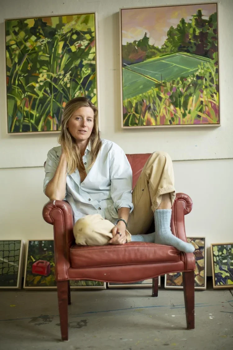 A woman sitting on a red vintage armchair in an art gallery, with one leg up and wearing light-colored pants and socks, holding a beige bag. Behind her, framed landscape paintings of greenery and tennis courts hang on the wall, with additional artwork leaning against the wall.