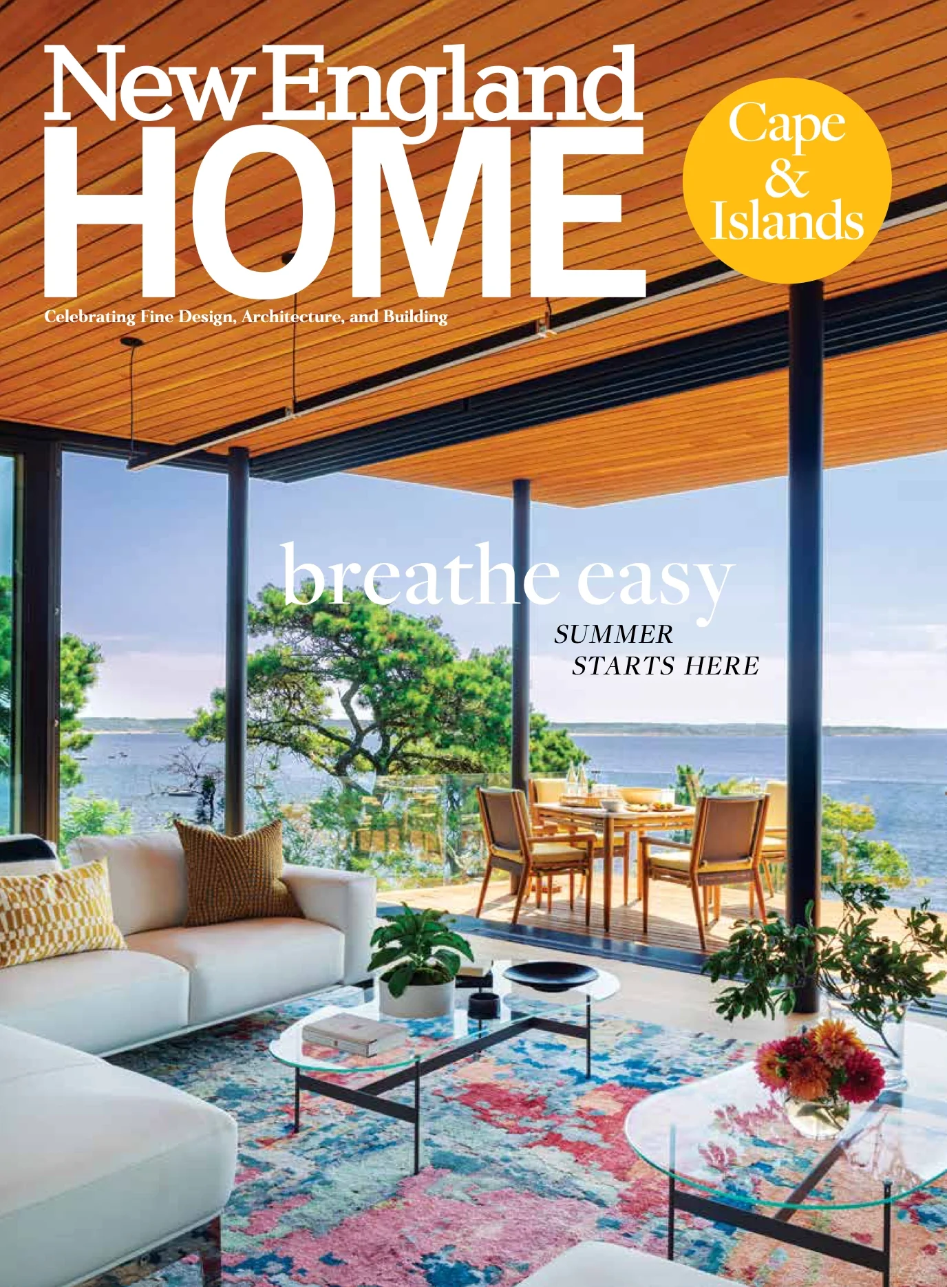 Interior of a modern living space with a view of a lake and trees, featuring a white sofa with yellow and brown cushions, a colorful rug, glass coffee tables with decor, a dining table with chairs on the patio, and a wooden ceiling.