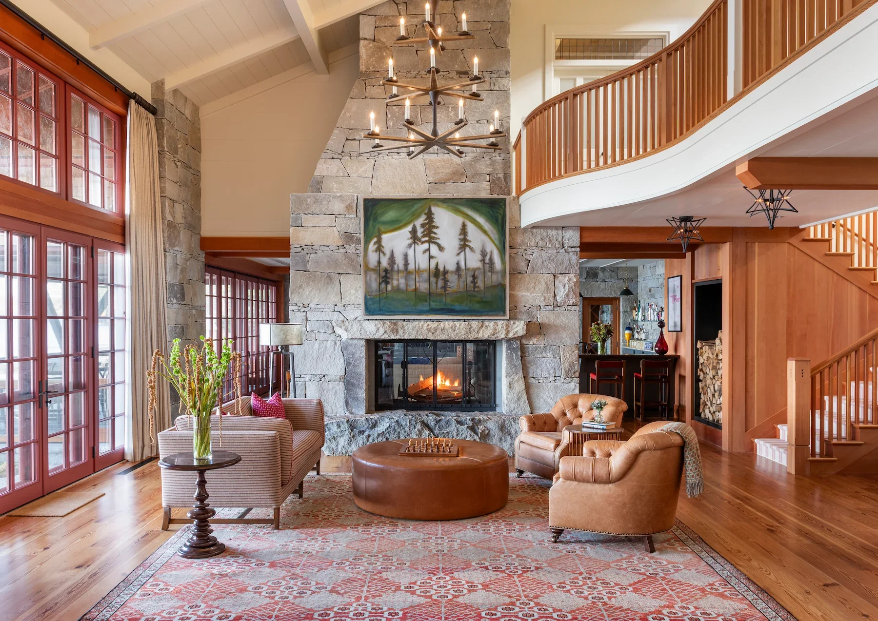 Living room with large stone fireplace, painting of trees above the fireplace, wooden ceiling and railing, leather armchairs, wooden coffee table, large windows with curtains, and a chandelier hanging from the ceiling.