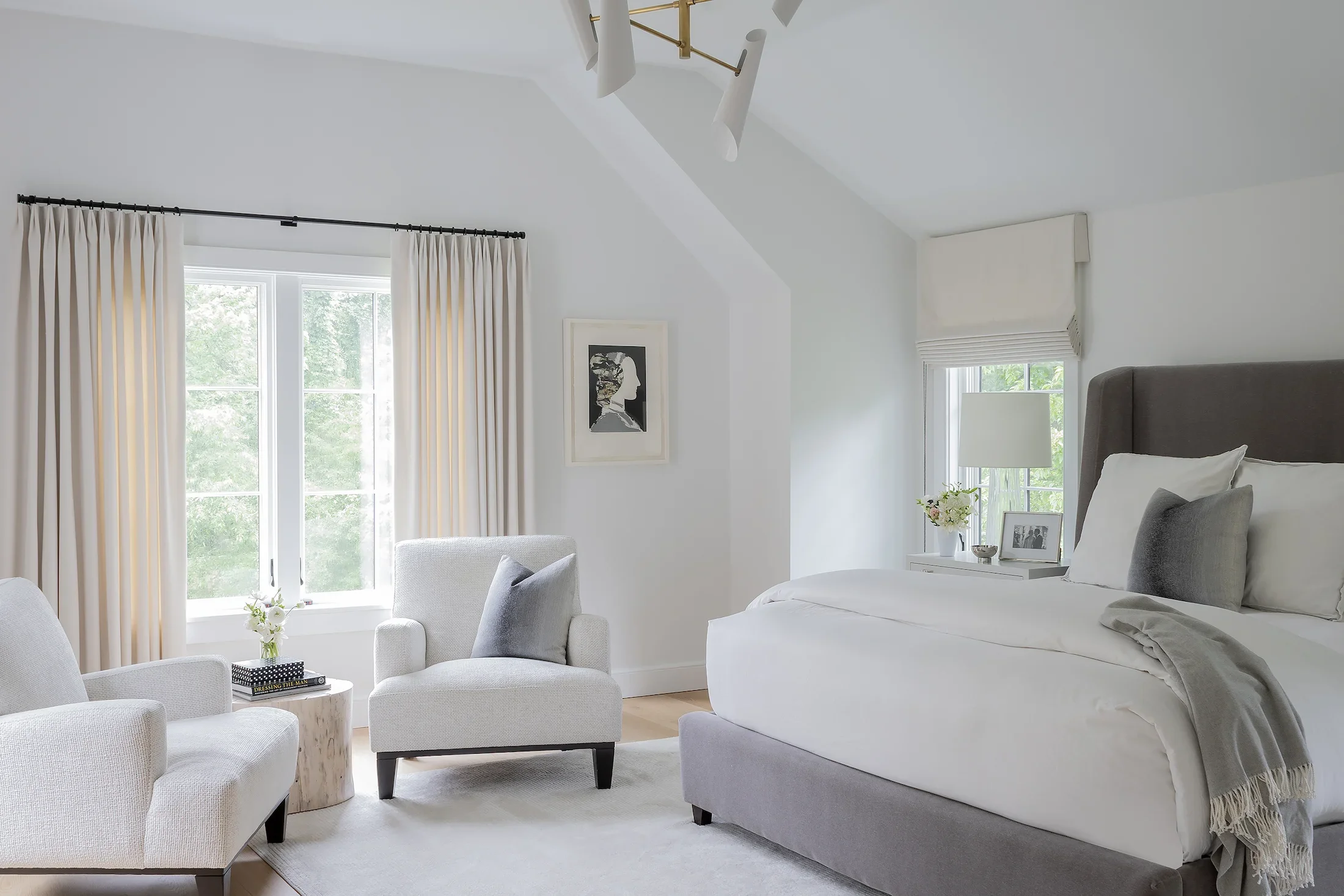 Bright bedroom with white walls, large window with cream curtains, gray upholstered bed, white nightstand with a lamp, framed photograph, and a vase of flowers, and a seating area with cream armchairs.