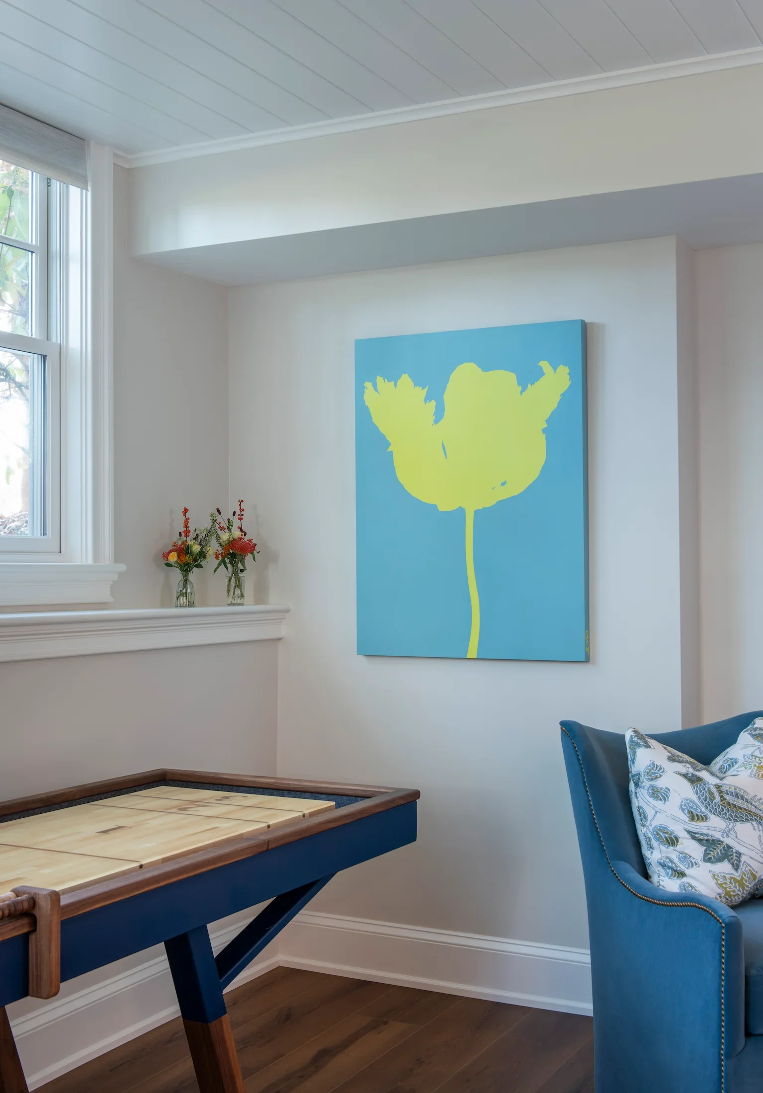 Corner of a living room with a window, white walls, a wooden floor, a blue chair with a patterned cushion, a wooden table, a painting of a blue background with a yellow-green flower, and two vases of flowers on a white shelf.
