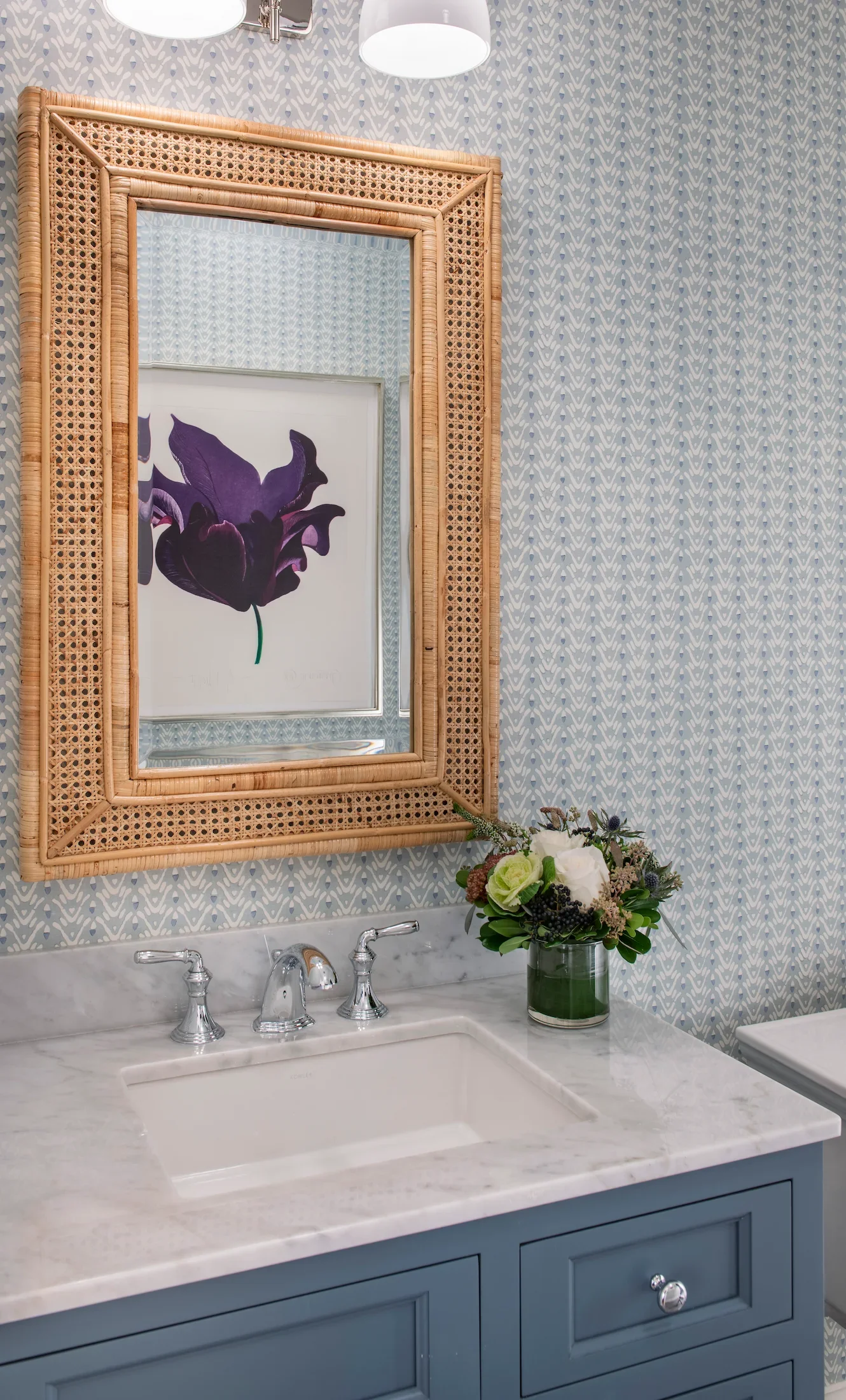 Bathroom sink with a white marble countertop, a blue cabinet with silver knobs, a woven framed mirror, a framed purple flower art, a floral arrangement in a green vase, patterned wallpaper, two white ceiling lights.