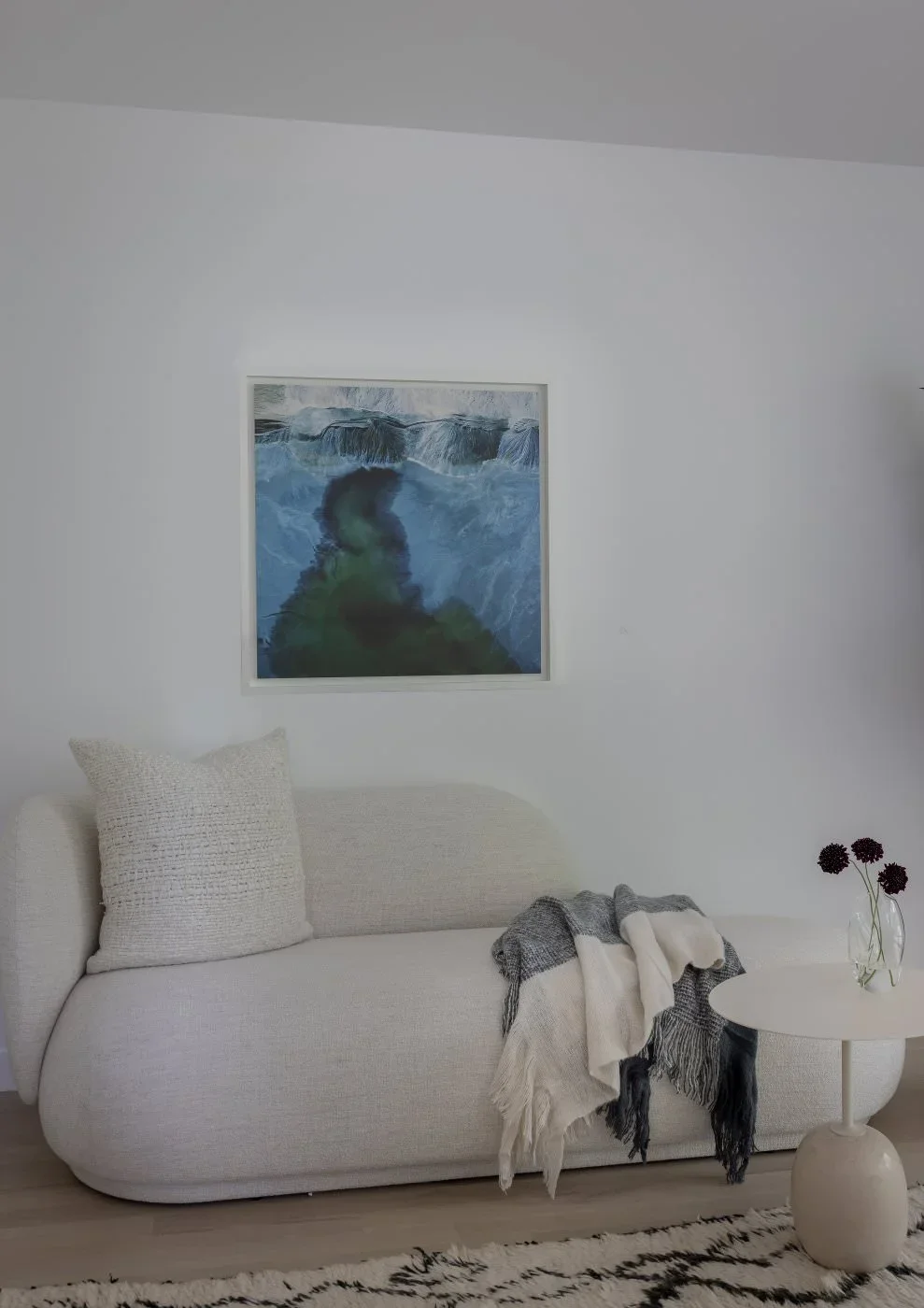 A minimalist living room with a white sofa, a light beige throw pillow, a black and white throw blanket, a small white round side table with a vase of dark purple flowers, a black and white rug, and a framed abstract artwork of a waterfall on the wall.