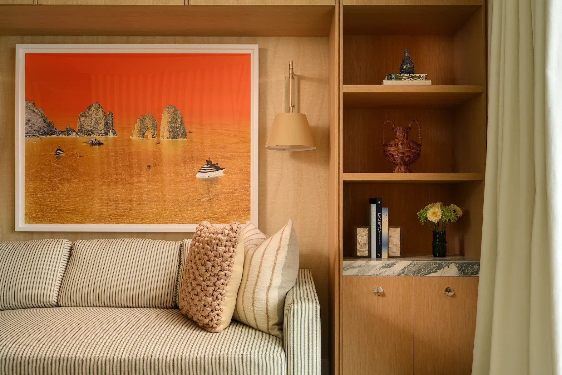 Living room with a striped sofa, decorative pillows, a large sunset landscape painting, a wall-mounted lamp, wooden shelving with decorative items, books, a flower vase, and cream-colored curtains.