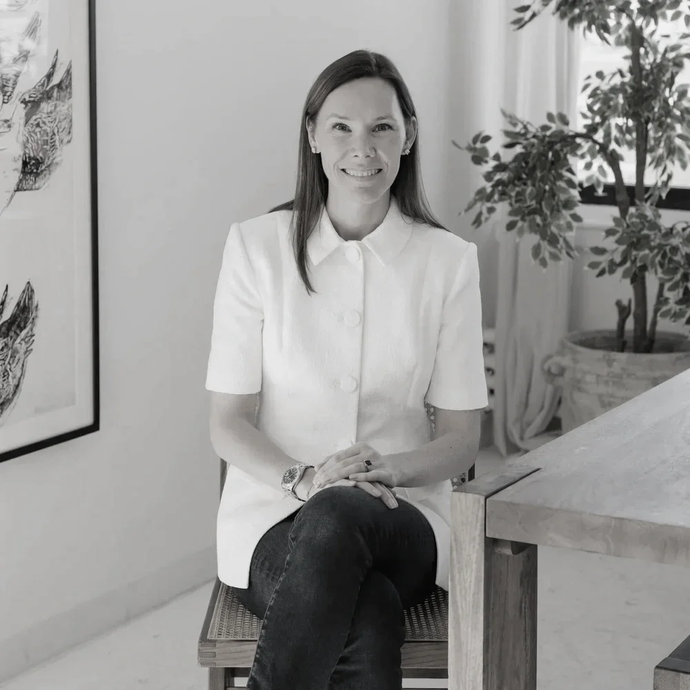 A woman with long hair, smiling, sitting on a chair at a table in a bright room with artwork on the wall and a potted plant nearby.