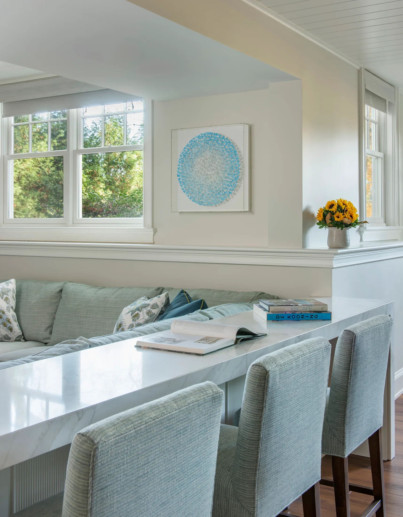 A bright room with large windows showing trees outside. There is a white marble table with books, a gray sofa with patterned pillows, a framed circular artwork with blue and white elements on the wall, and a vase of yellow sunflowers in the corner.