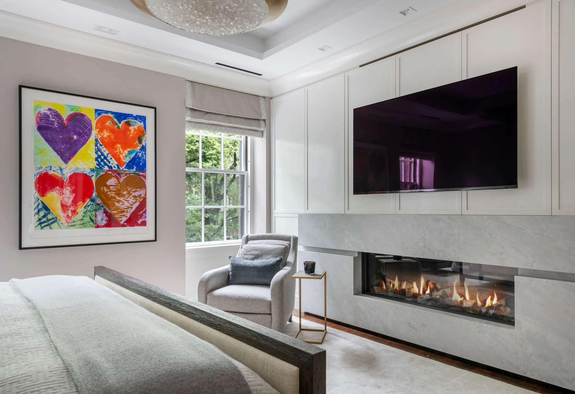 Modern living room with a painting of colorful hearts on the wall, a window with a view of greenery, a fireplace, and a gray armchair.