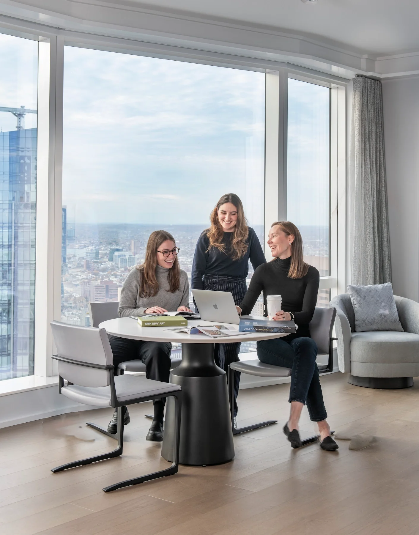 Three women having a discussion in a modern office with a city view. Two are seated at a round table with books, a magazine, and a laptop. One woman stands, smiling, with a coffee cup in hand.