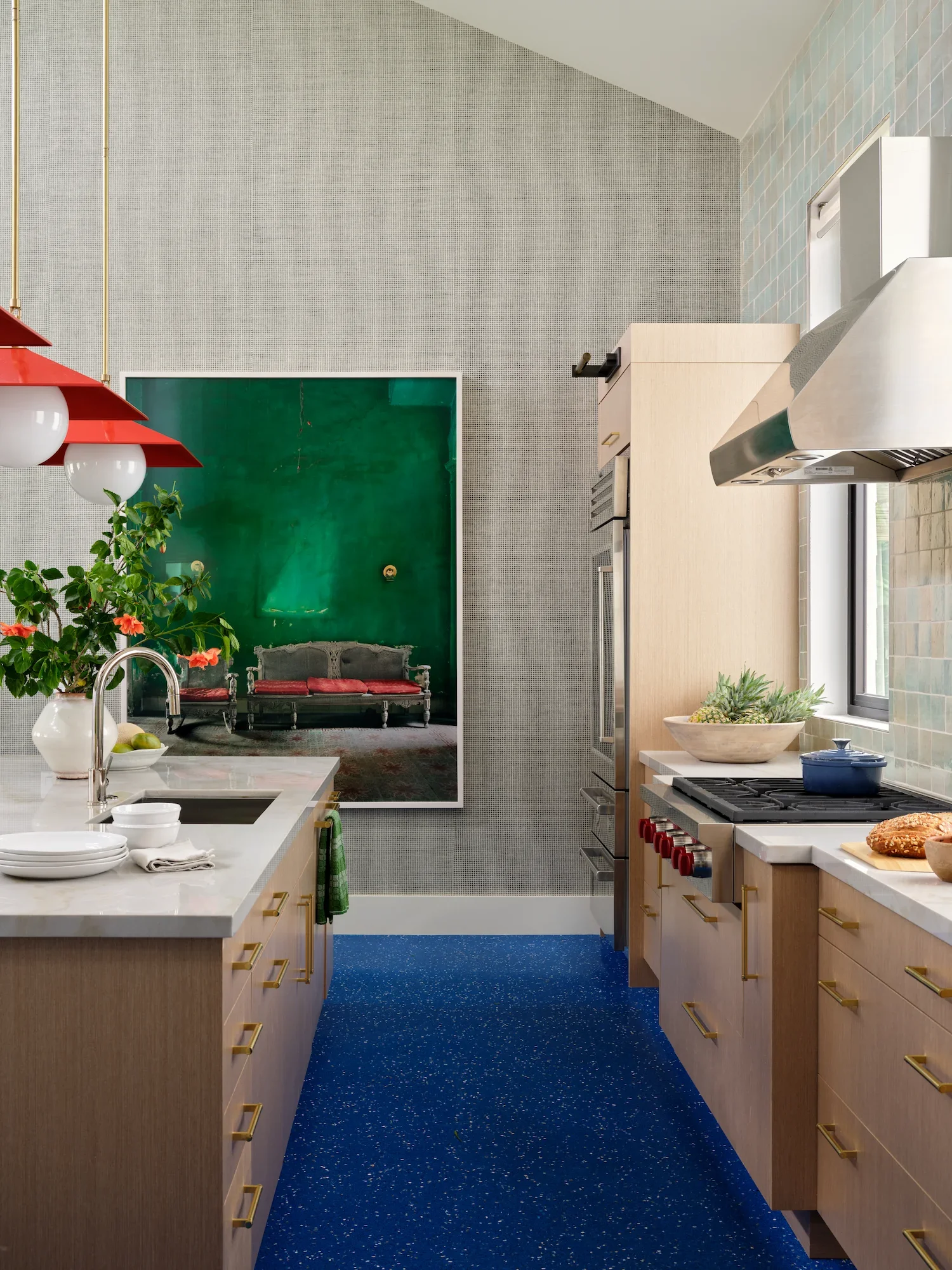 A modern kitchen with light wood cabinets, a white marble island, red pendant lights, green plants, a blue speckled carpet, a window, and a green framed mirror. There is also a seating area with a vintage sofa and a large green wall.