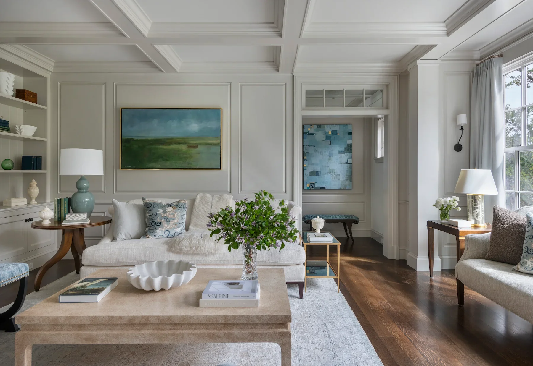 A beautifully decorated living room with white paneled walls and coffered ceiling, featuring a white sofa with decorative pillows, a wooden side table with a large ceramic vase lamp, framed landscape and abstract paintings, floral arrangements, a coffee table with books and a white decorative bowl, a window with white curtains, and additional seating with decorative pillows.