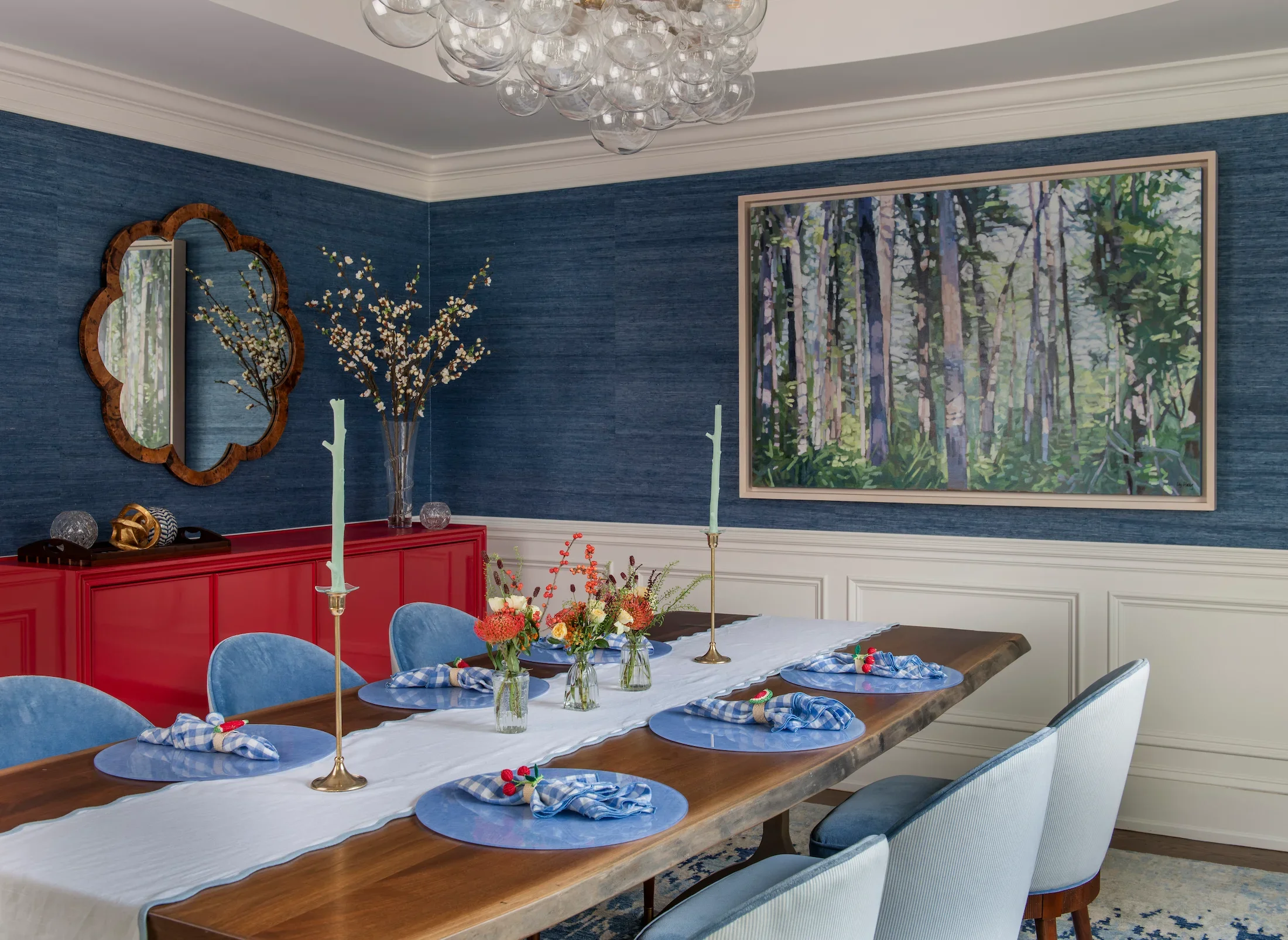 A dining room with a long wooden table set with blue placemats, napkins with checkered pattern and small decorations. The room has blue walls, white crown molding, and a large painting of a forest on the wall. There is a red sideboard with decorative items, a mirror with a wooden frame, and a table centerpiece with flowers and two tall, light green candlesticks.