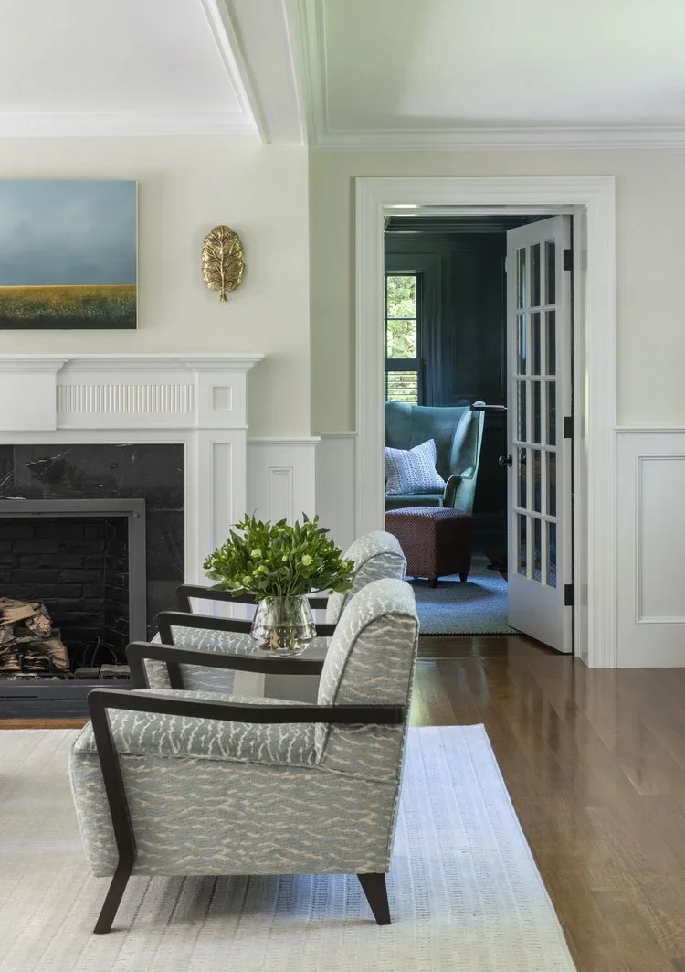 Living room with a fireplace, three patterned armchairs, a glass vase with green flowers, and an open door leading to a cozy sitting area with a blue armchair and window.