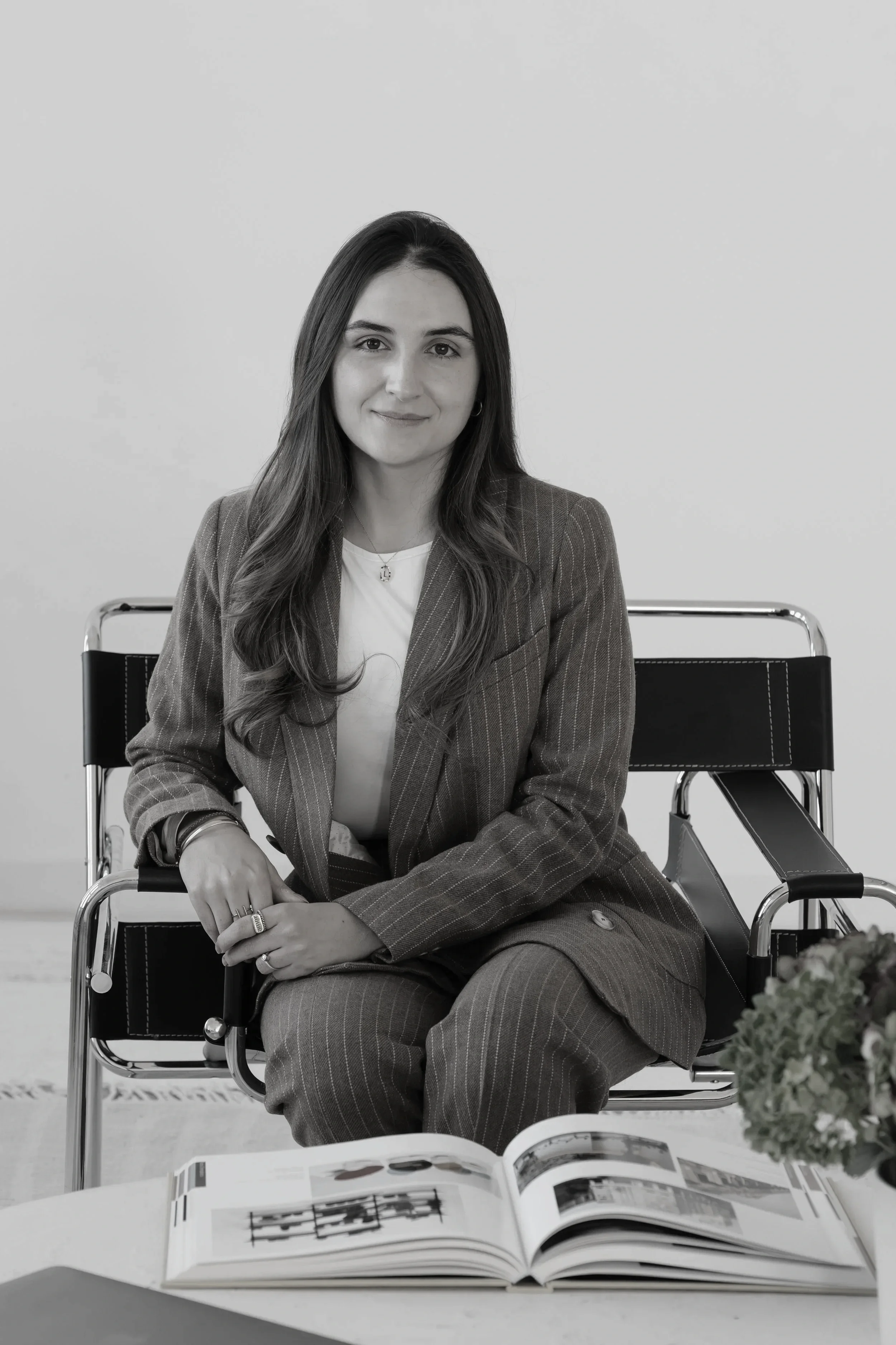 A black and white photo of a woman with long hair sitting in a chair at a table with an open magazine in front of her. She is wearing a pinstripe suit and a necklace, looking at the camera with a slight smile. There is a bouquet of flowers on the table to the right.