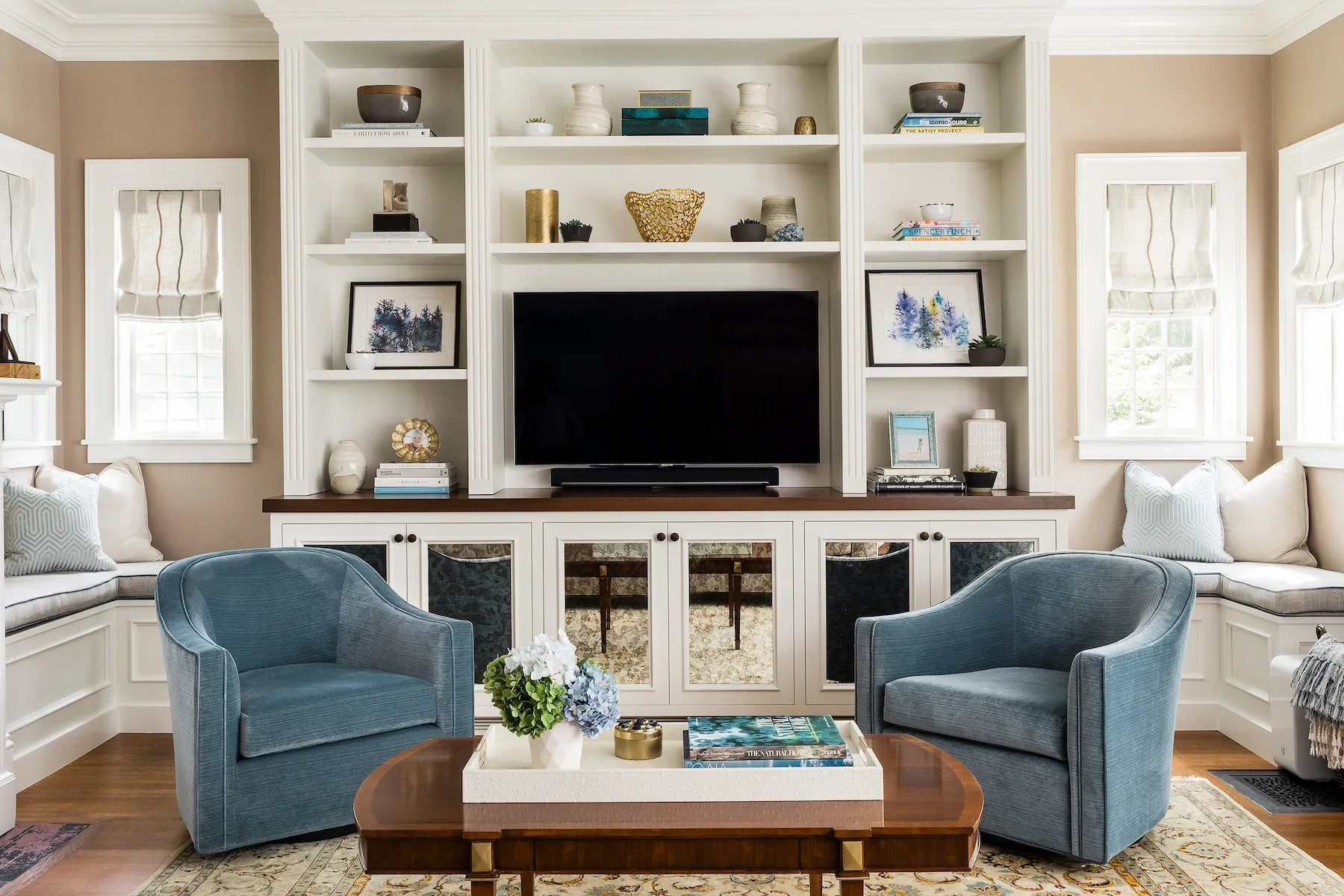 Living room with built-in white shelving unit above a television, surrounded by windows with white covers, beige walls, and light-colored furniture including dark blue armchairs and a wooden coffee table with flowers and books.