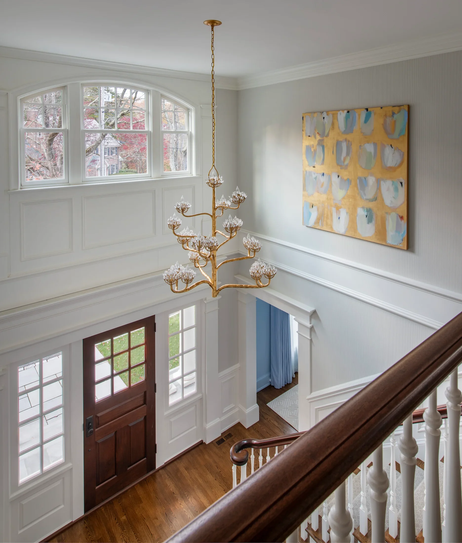 Entryway of a house featuring a wooden front door with glass panes, a large window with multiple sections and a curved top, a grand staircase with a wooden handrail and white balusters, warm hardwood flooring, white walls with decorative molding, a large abstract painting with blue, white, gray, and gold colors, and a gold chandelier hanging from the ceiling.