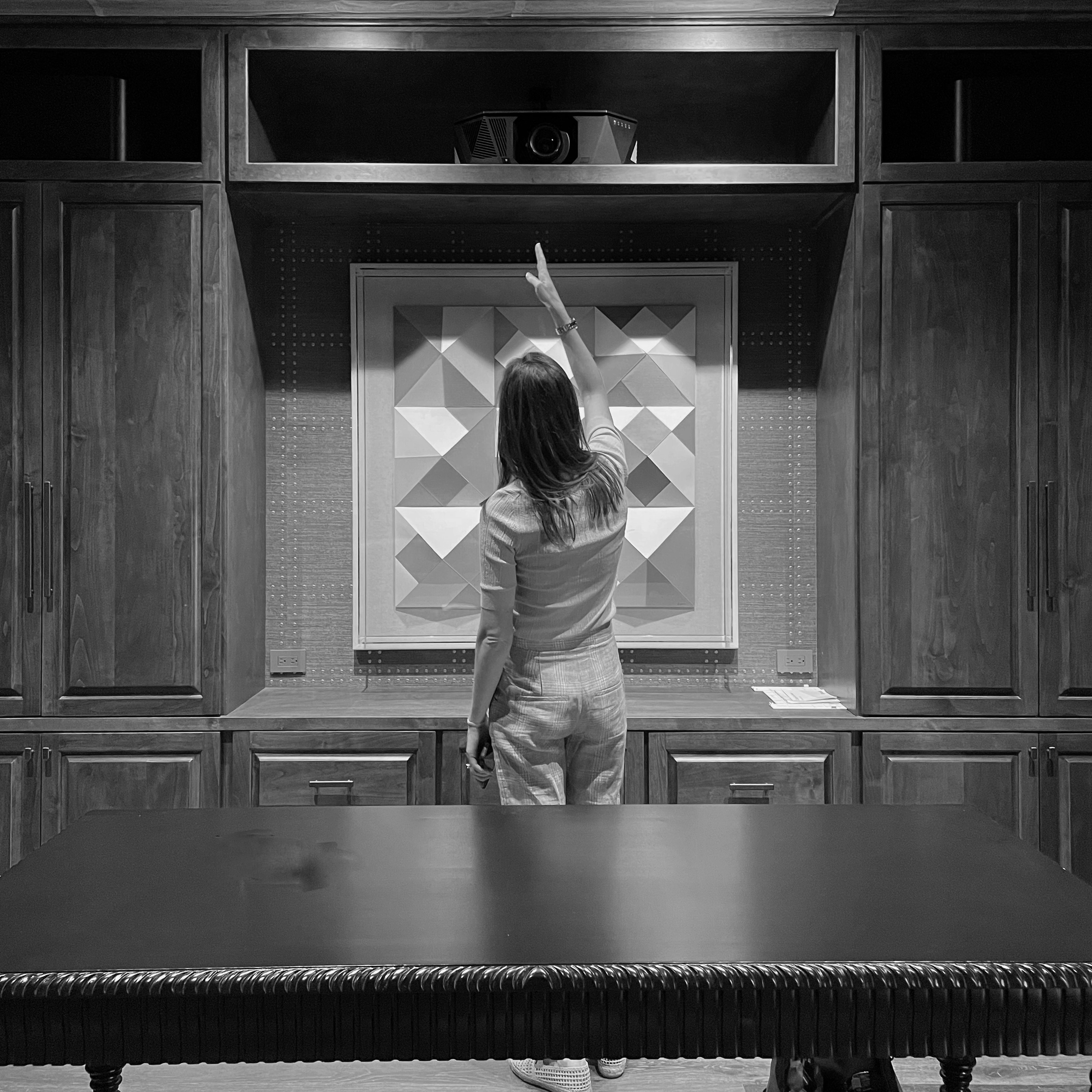 A woman with long hair reaching up to touch a projector mounted on a black wooden cabinet in a room with decorative wall panels and a dark wooden table with a textured edge.