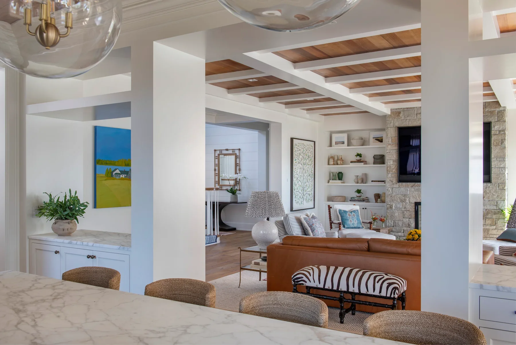A bright and airy living room with a white and wood ceiling, a stone fireplace, a wall-mounted TV, white built-in shelves with decor, a brown leather sofa, a zebra-print ottoman, and a mix of patterned pillows.