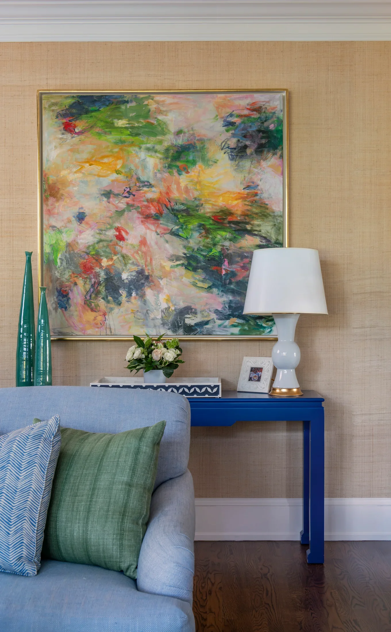 Living room corner with beige walls, a large abstract painting with green, pink, and orange colors, a white table lamp on a blue console table, a small floral arrangement, and pillows on a light grey sofa.