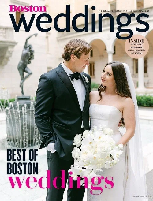 A bride and groom in wedding attire standing outdoors in front of a fountain, looking at each other, with the groom in a tuxedo and the bride holding a bouquet of white flowers.