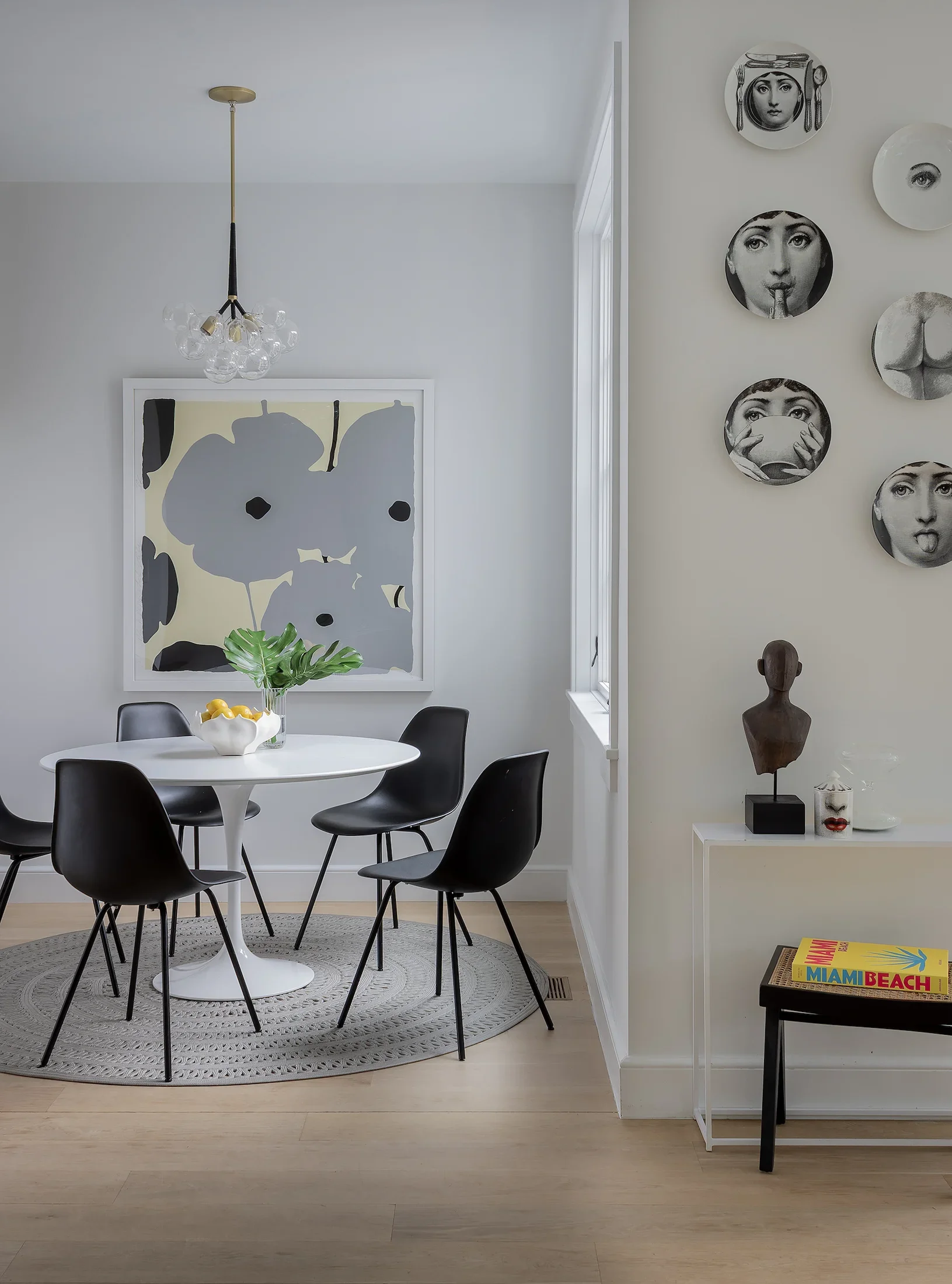 A dining room with a white round table surrounded by six black chairs, a woven rug, a framed minimalist art piece, a chandelier, a potted plant, and decorative wall plates with black and white faces, plus a small table with a "Miami Beach" magazine and sculptures.