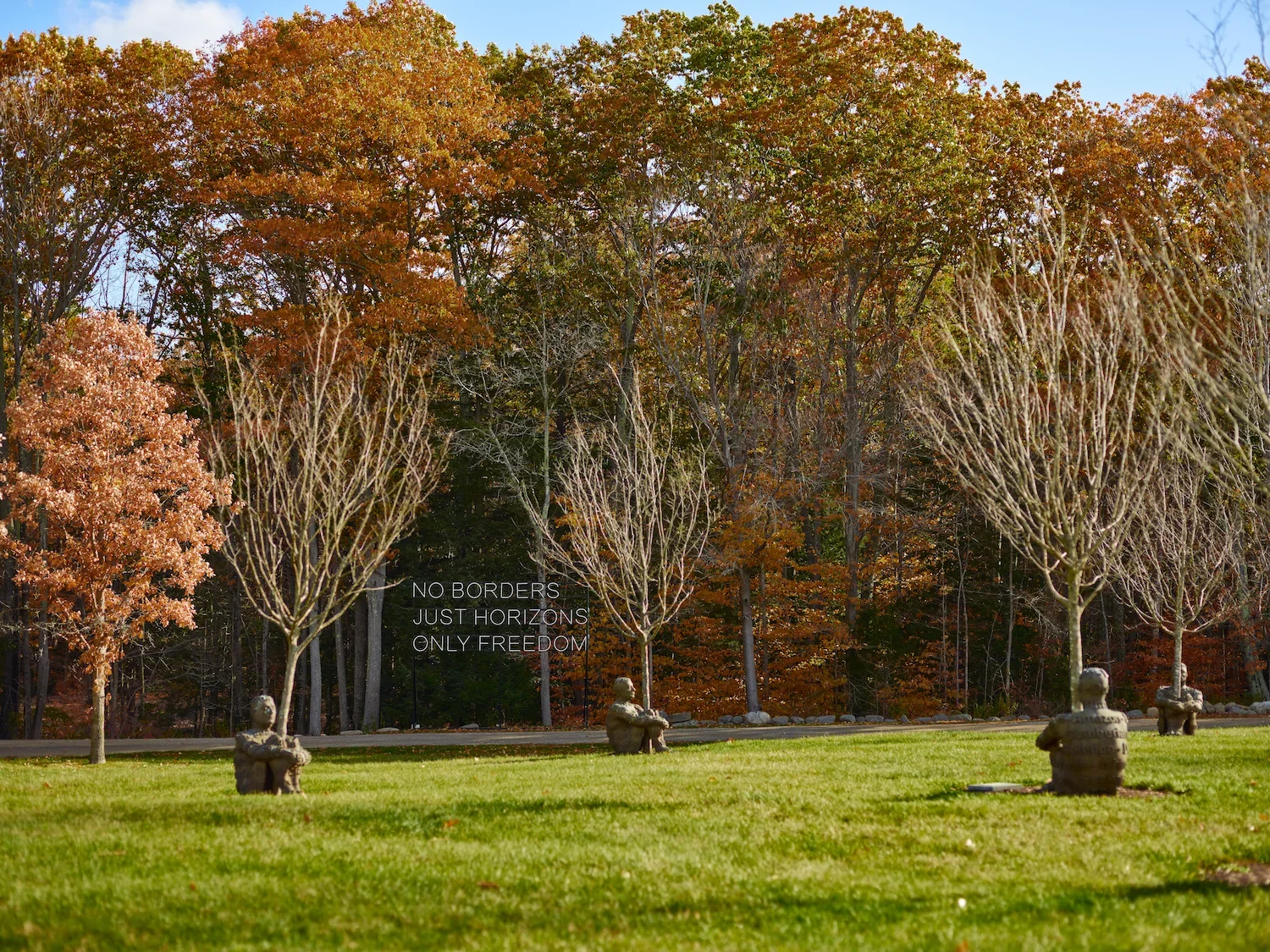 A park with four bronze sculptures of children sitting on the grass, a line of trees with autumn foliage in the background, and a black sign with white text that reads: 'NO BORDERS, JUST HORIZONS, ONLY FREEDOM'.