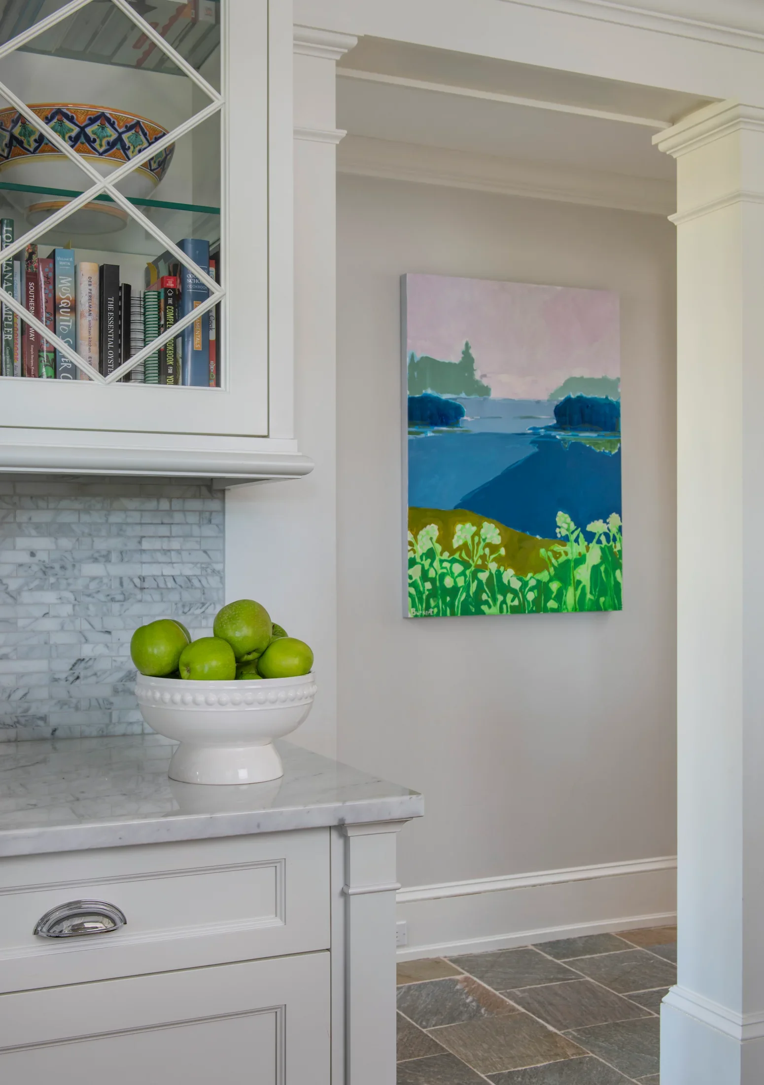 A white ceramic bowl filled with green apples on a marble countertop in a bright kitchen. Part of a white cabinetry with glass doors showing a bowl with colorful geometric patterns and a collection of books inside. An abstract landscape painting with a body of water, trees, and a pink sky on a wall in the background, and stone flooring.