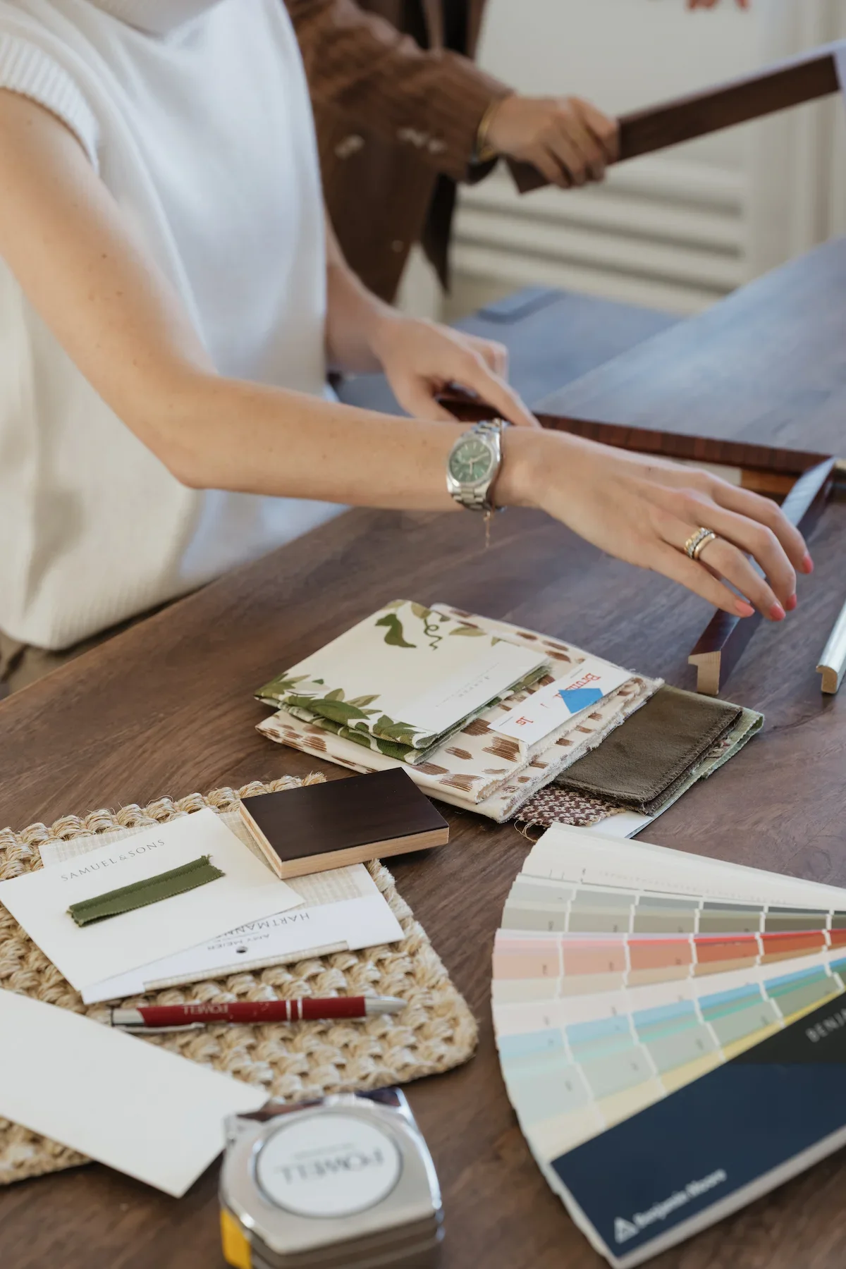 A person with a silver watch on their left wrist and a wedding ring on their left hand, sitting at a wooden table. The table has fabric swatches, color palette samples, and design catalogs for interior decorating or sewing. A second person in the background holds a wooden plank.